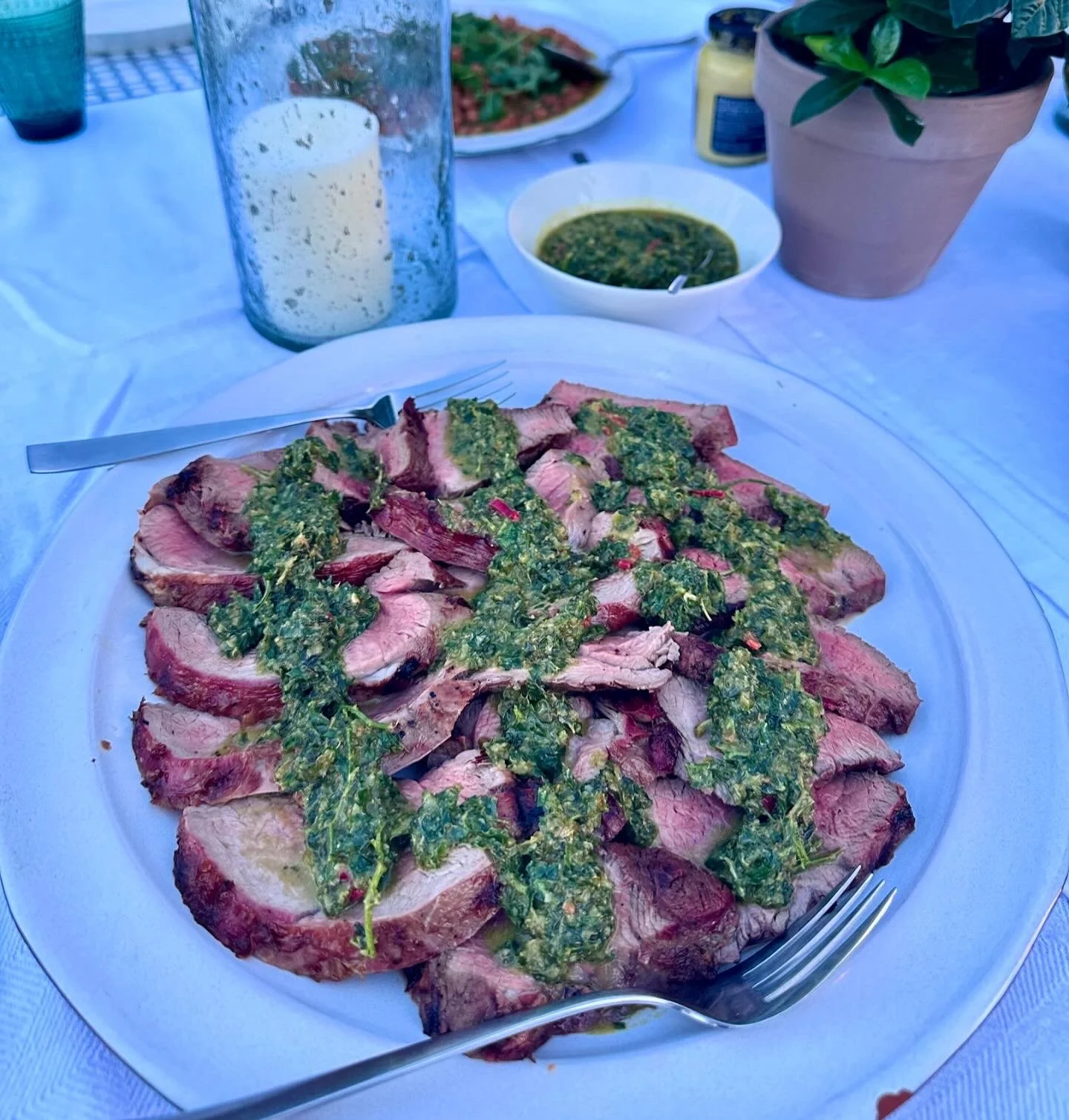 Thinly sliced medium-rare beef steak topped with green herb sauce on a white plate, with a fork and knife.