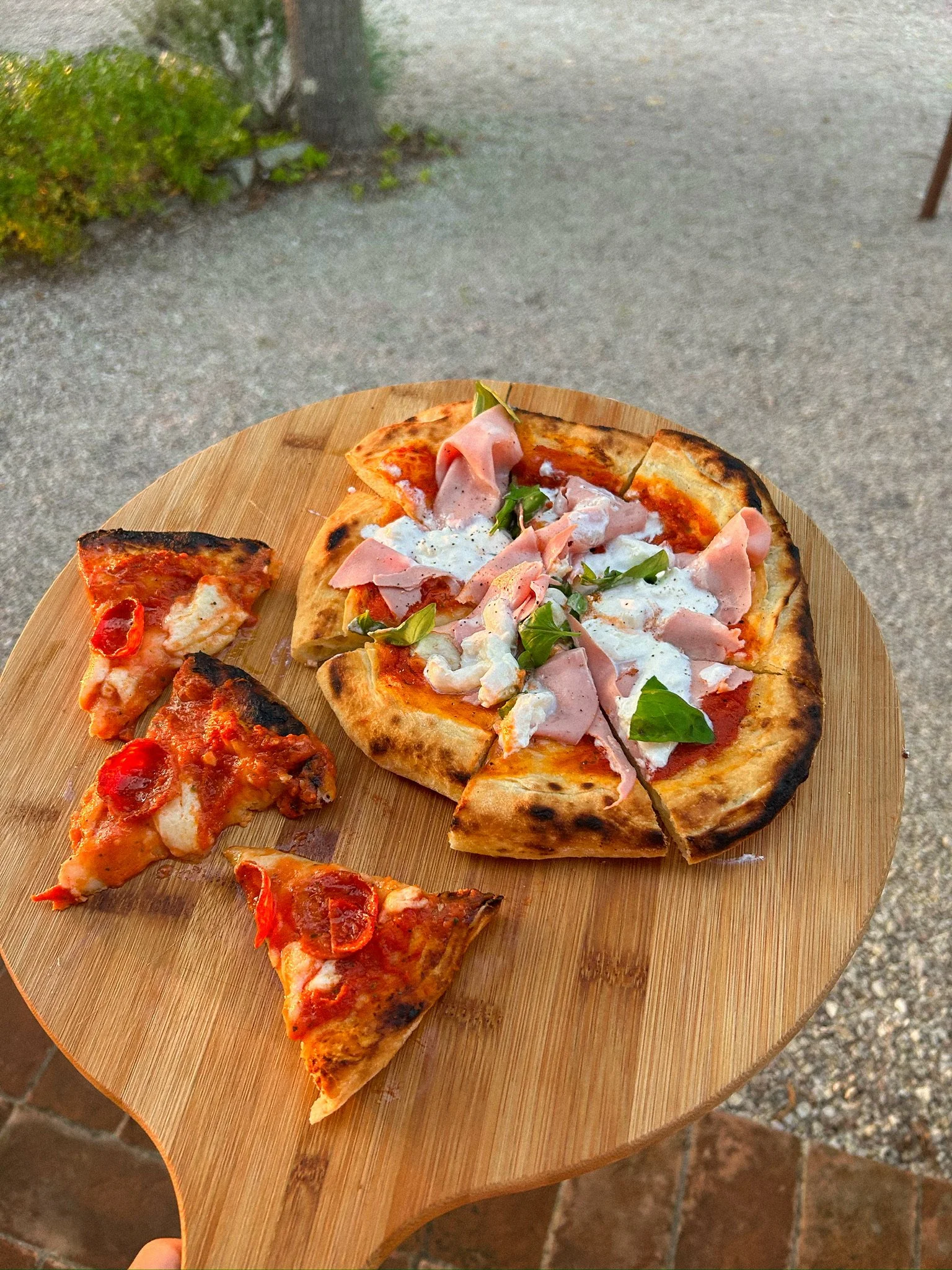 A wooden pizza peel holding a slice of pepperoni pizza and a whole pizza topped with ham, cheese, and basil, outdoors on a stone patio.