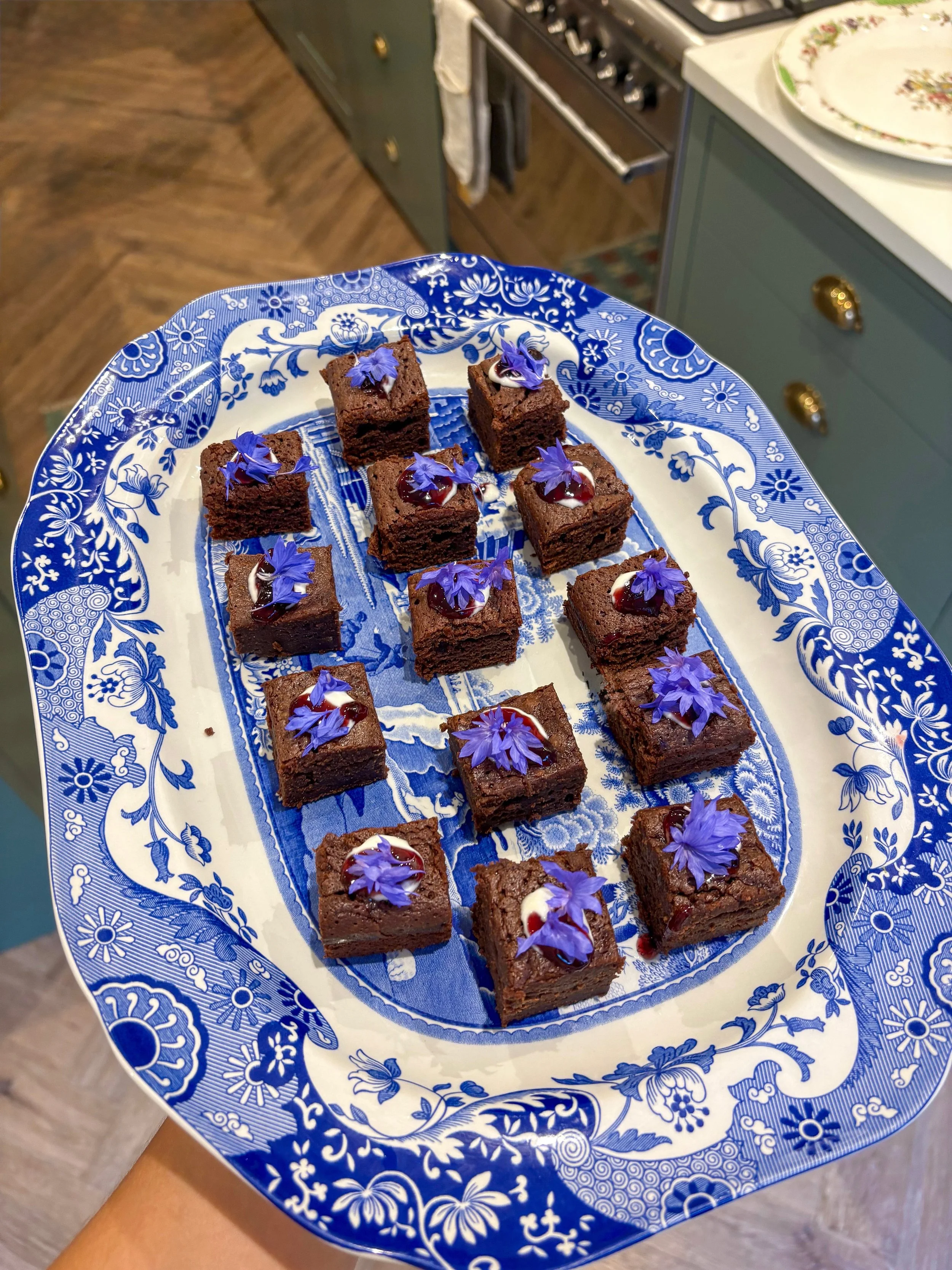 A blue and white patterned plate with small chocolate brownies topped with purple edible flowers and drizzled with white and red syrup, being held in a kitchen.