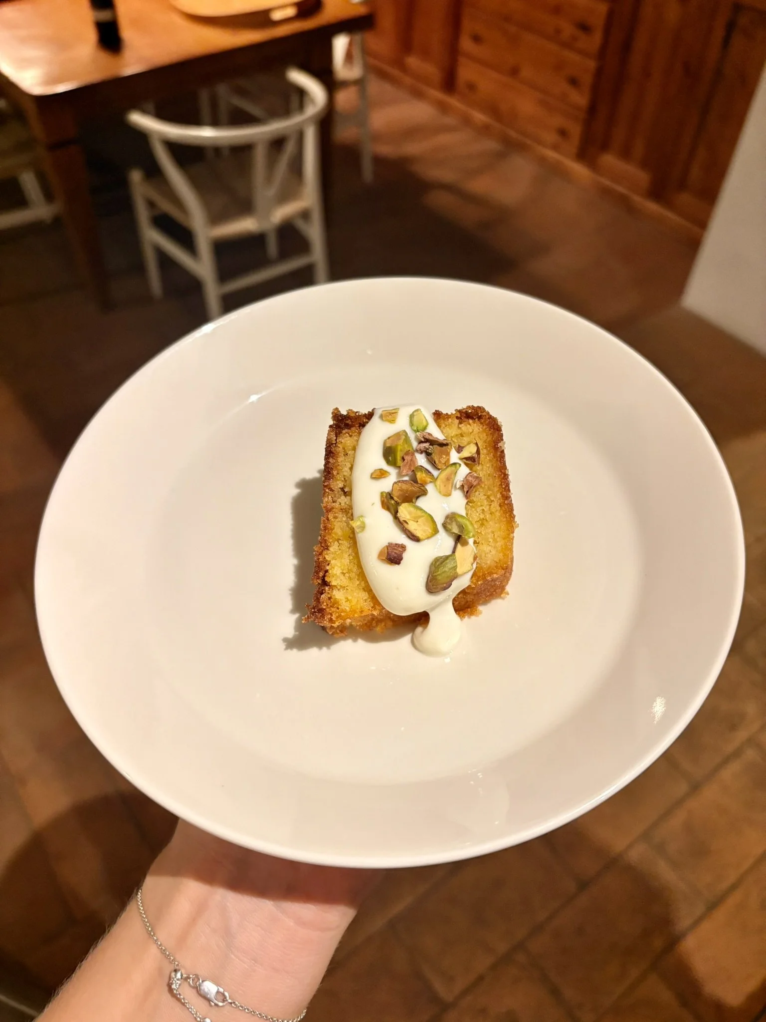 A slice of cake topped with whipped cream and chopped pistachios on a white plate, held by a person wearing a silver bracelet, in a wooden-paneled room.