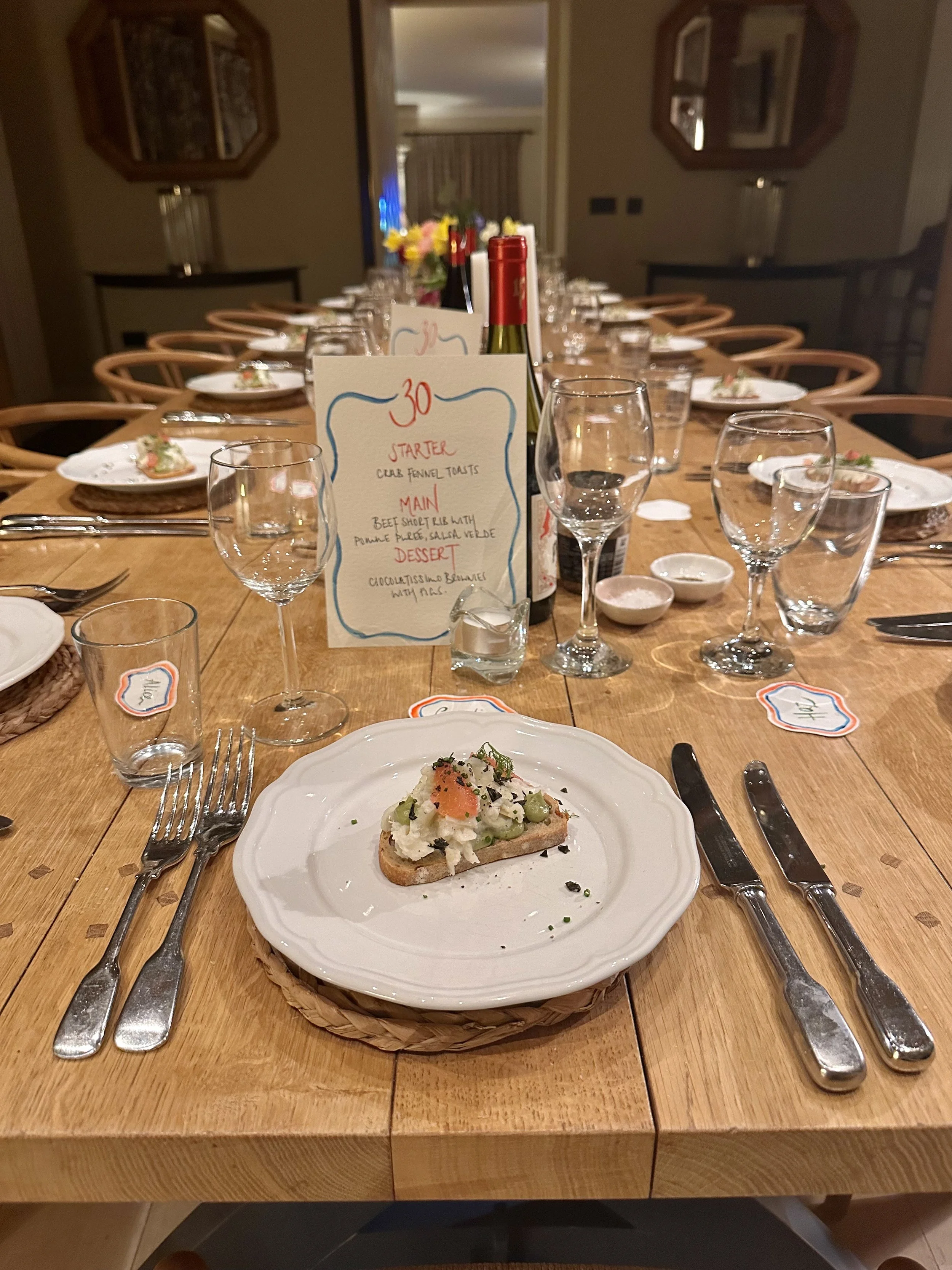 A long dining table set for a meal with plates, glasses, utensils, and bottles of wine. A small piece of bread with toppings is in the foreground. A menu card is in the center of the table with a list of starter, main, and dessert options.
