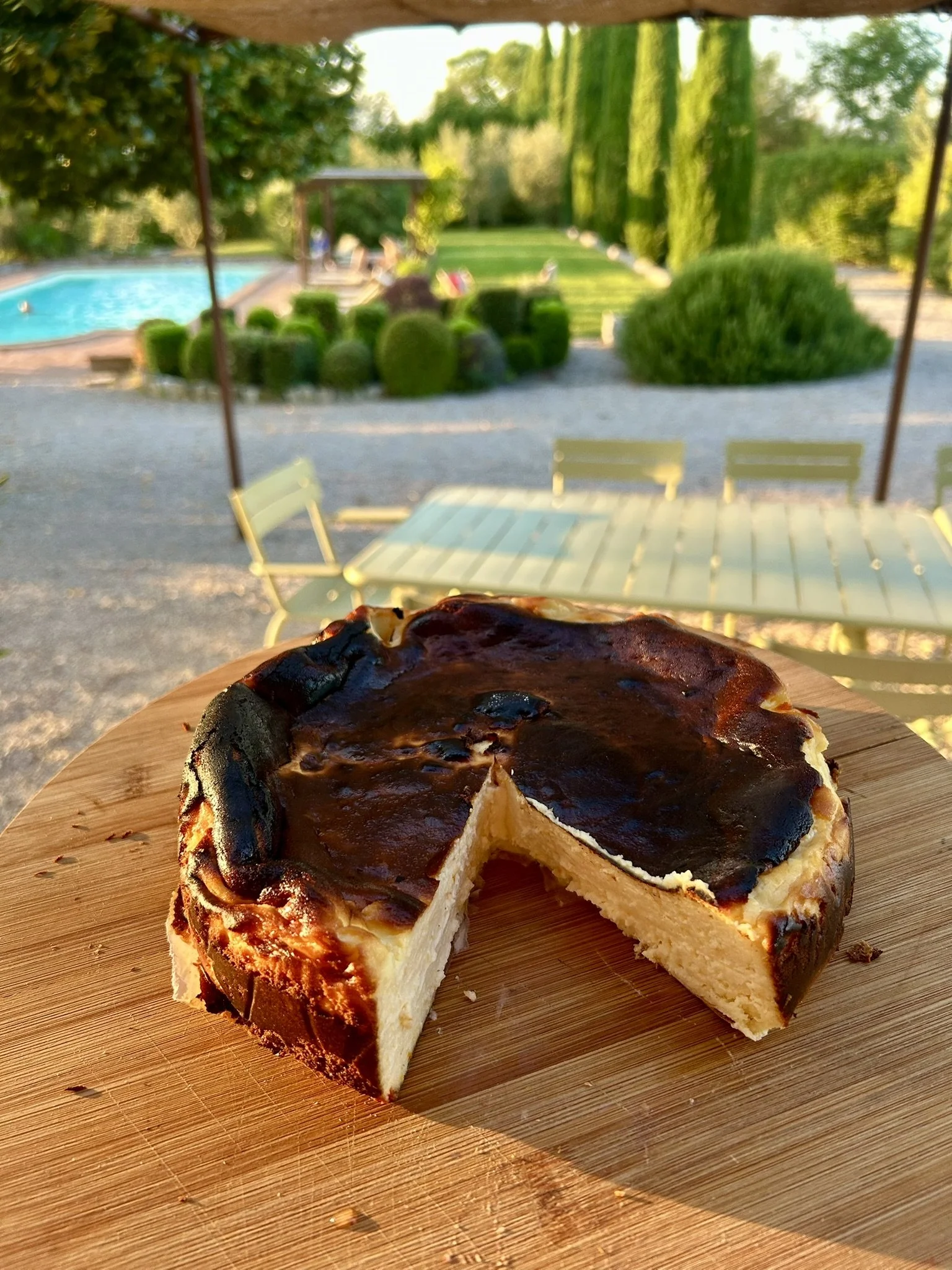 A cheesecake with a burnt top and a slice removed on a wooden cutting board, with a backyard pool and garden in the background.