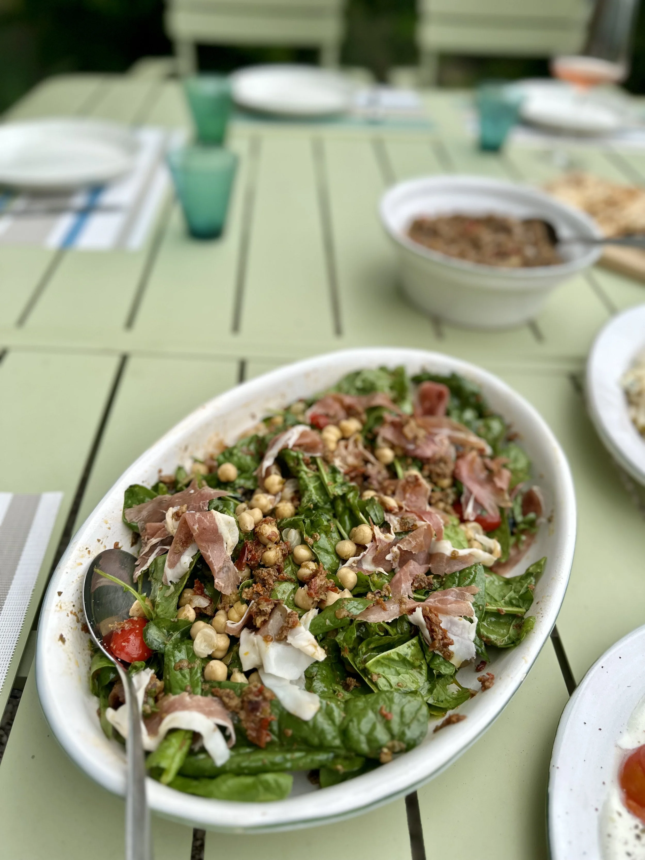 A fresh spinach salad with cherry tomatoes, bacon, nuts, and cheese on a light green outdoor table.