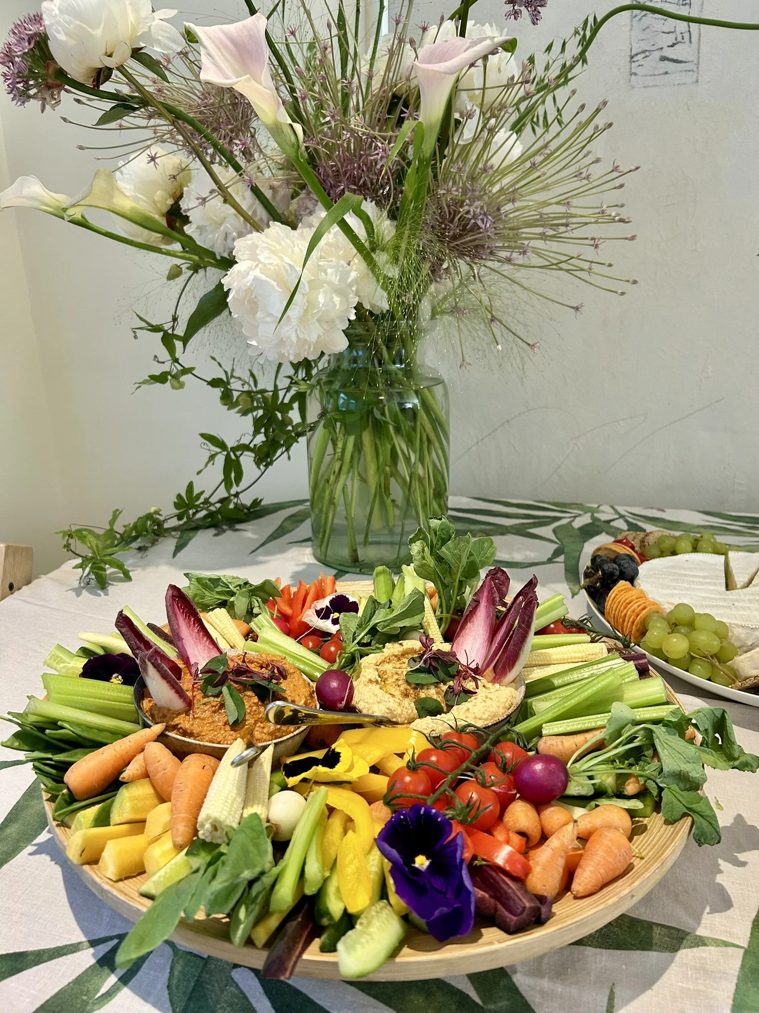 A large wooden platter filled with assorted fresh vegetables, including carrots, cherry tomatoes, celery, yellow peppers, radicchio, and edible flowers. In the background, there's a glass vase with a bouquet of white, pink, and purple flowers on a ta