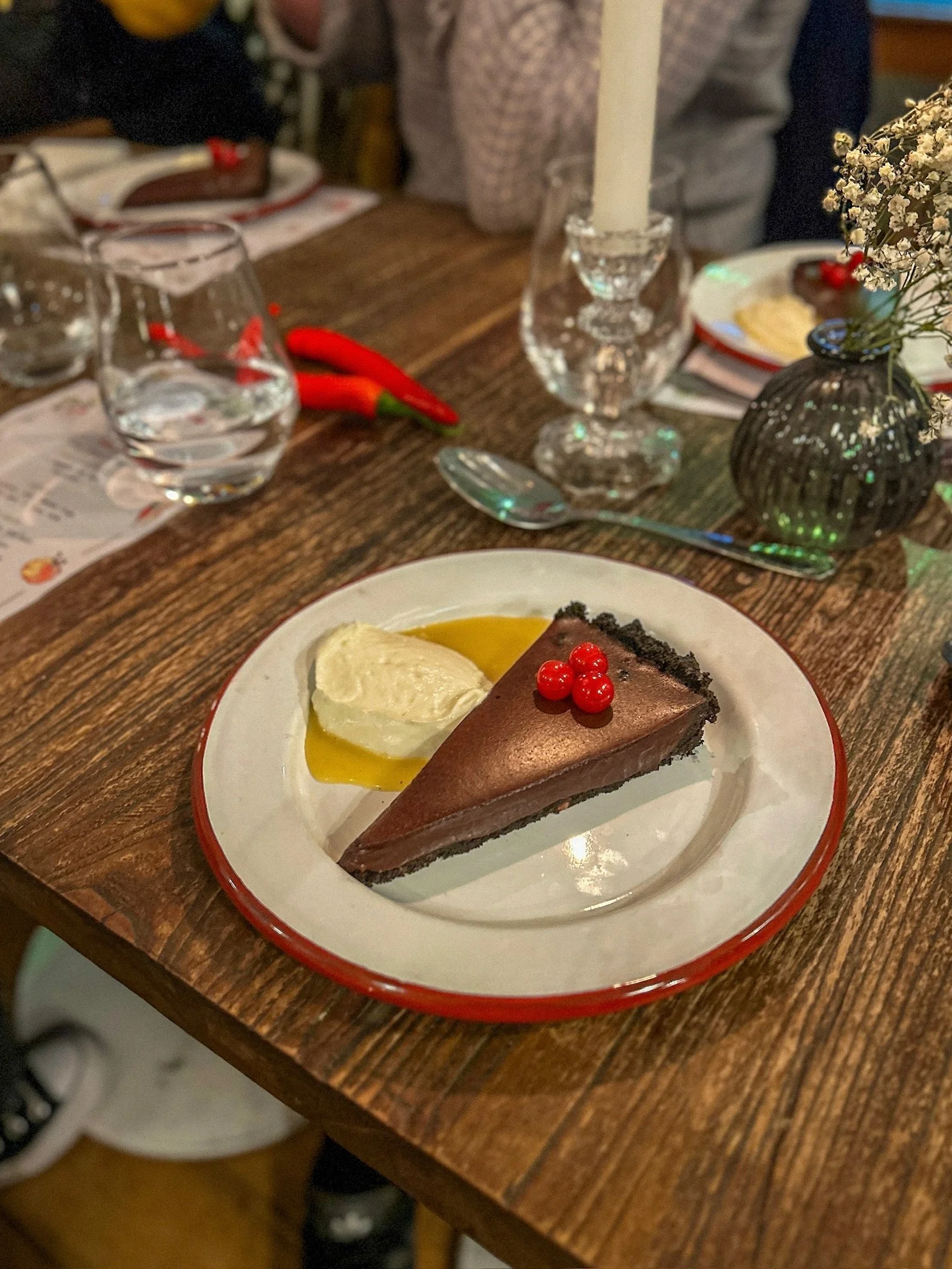 Chocolate cake with red berries on top, served with a dollop of whipped cream and a drizzle of honey, on a white plate with a red rim, on a wooden table.