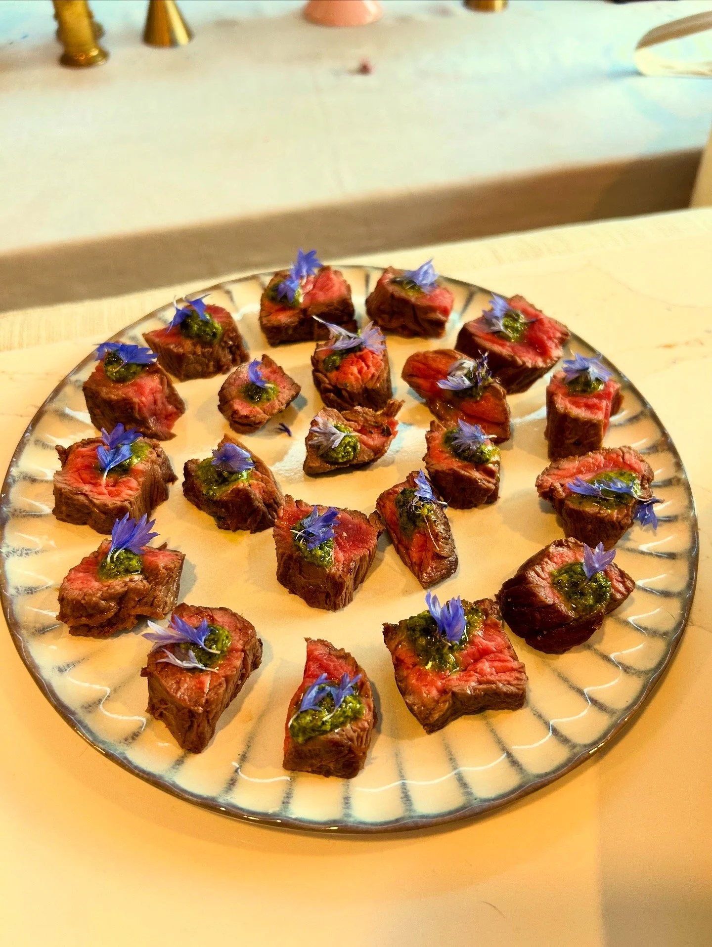 Close-up of small, cooked pieces of steak topped with green sauce and edible purple flowers on a decorative ceramic plate.