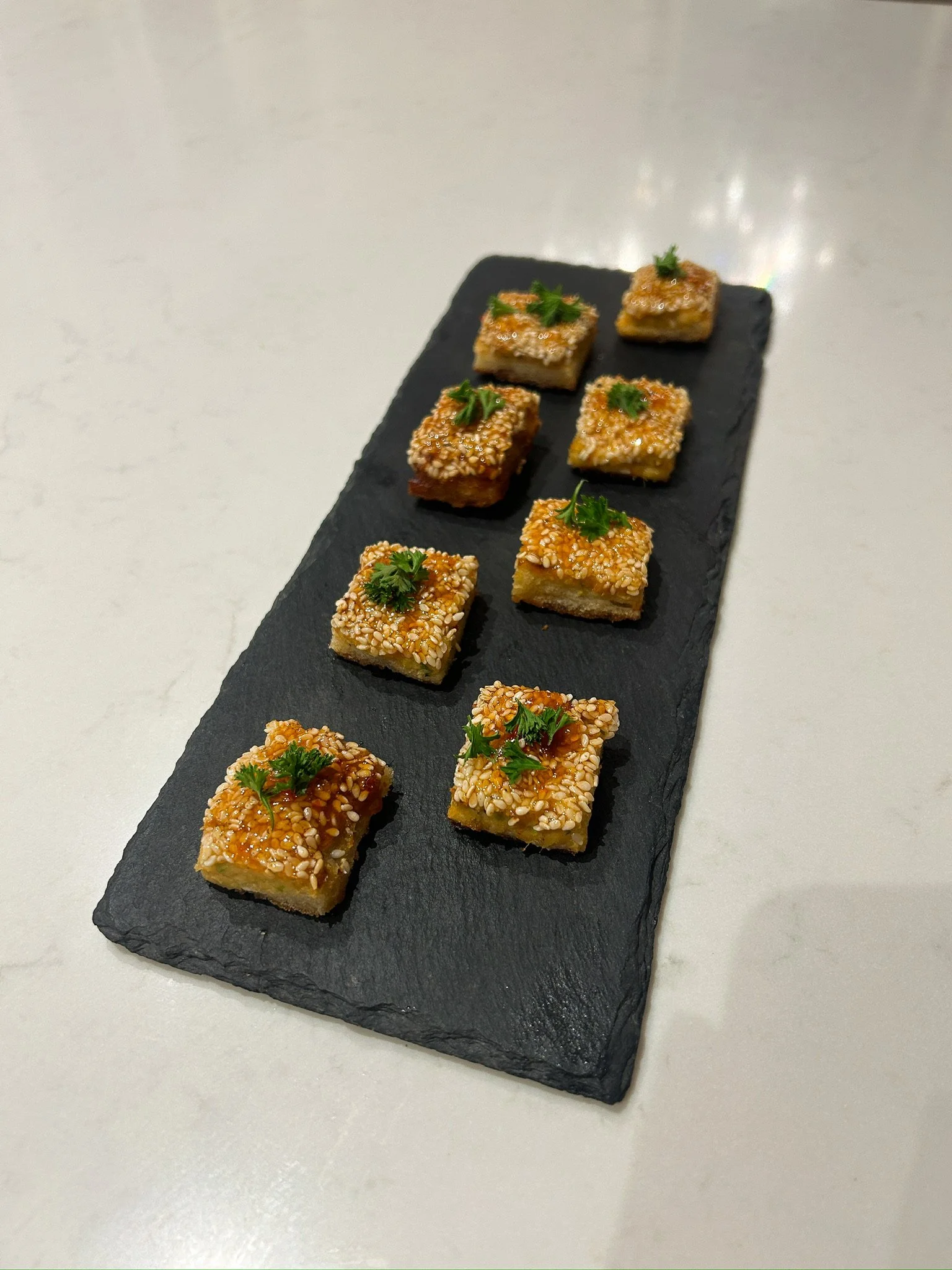 Nine small square appetizers topped with sesame seeds and garnished with parsley, arranged on a black slate serving platter.