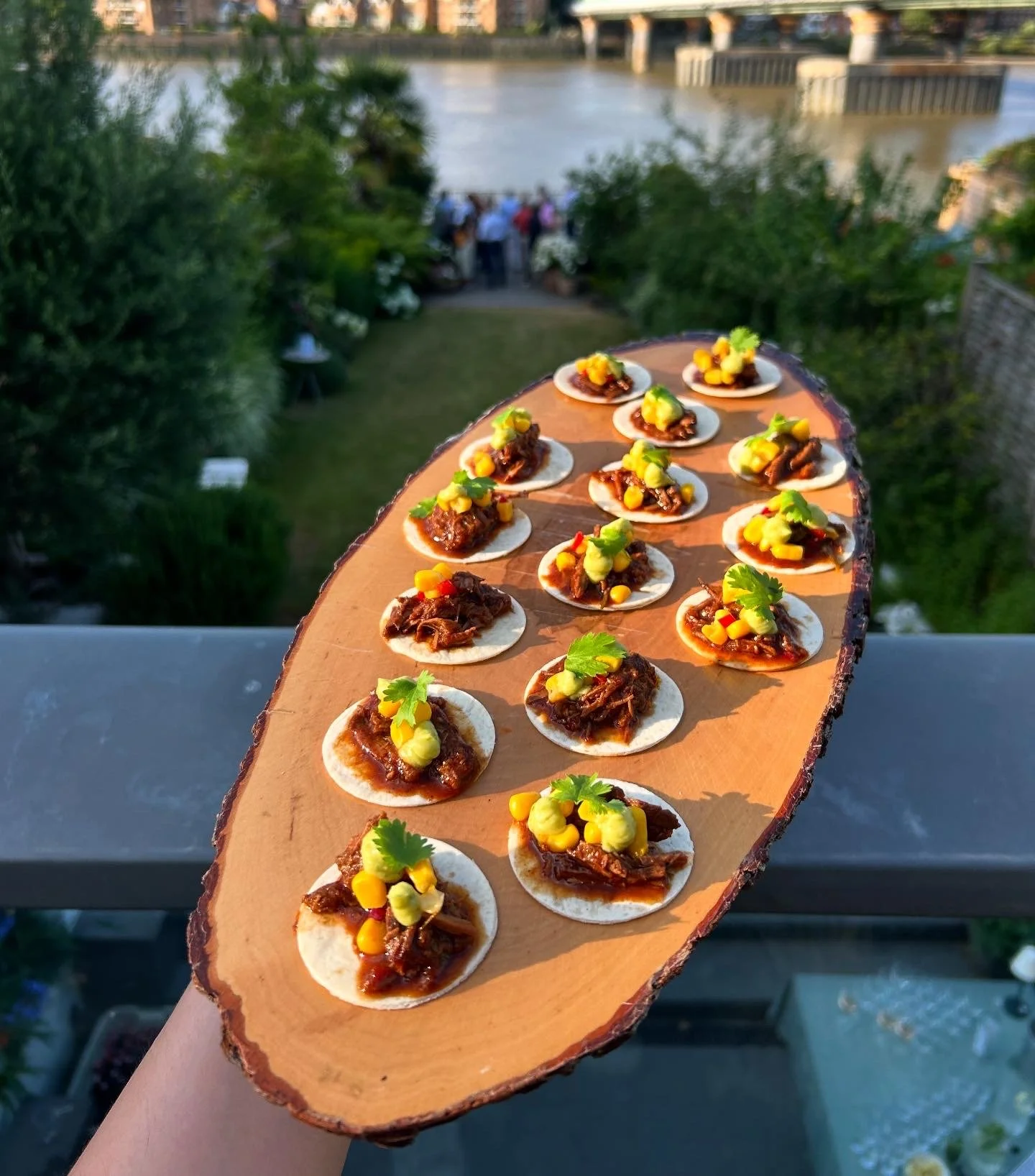 A person is holding a wooden platter of small appetizers topped with meat, yellow sauce, and green herbs, with a riverside outdoor gathering in the background.