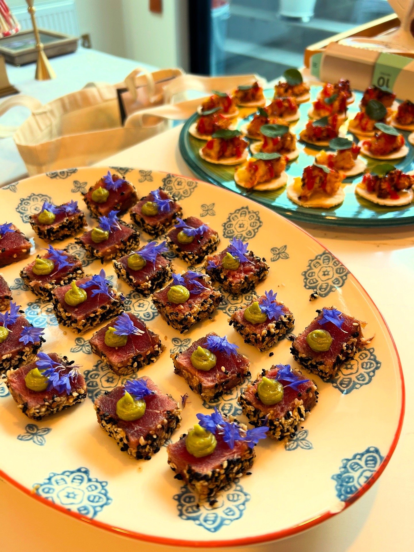 Two plates of small, decorated appetizers on a table, with one plate featuring seared tuna slices topped with green sauce and blue edible flowers, and the other with mini tacos topped with green leaves.