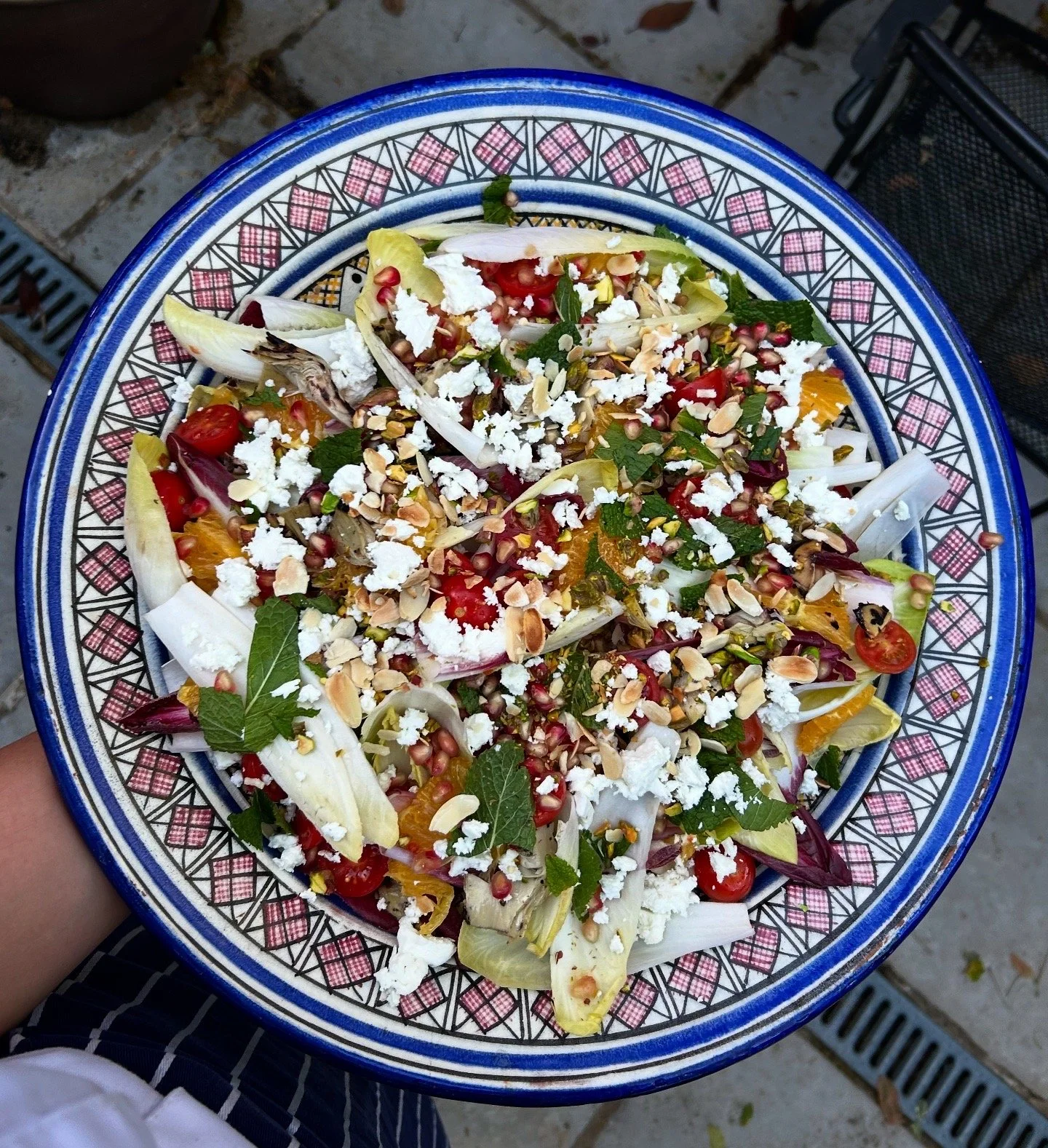 Colorful salad on a decorative blue and white plate, featuring cherry tomatoes, leafy greens, sliced vegetables, sprinkled cheese, pomegranate seeds, sliced almonds, and herbs.