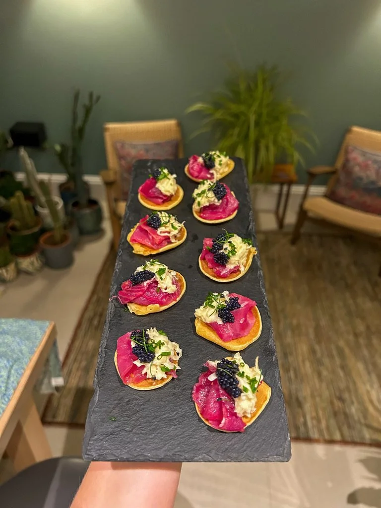 Appetizer bites with pink, cream, and blackberries on a slate platter, held indoors with chairs and plants in the background.