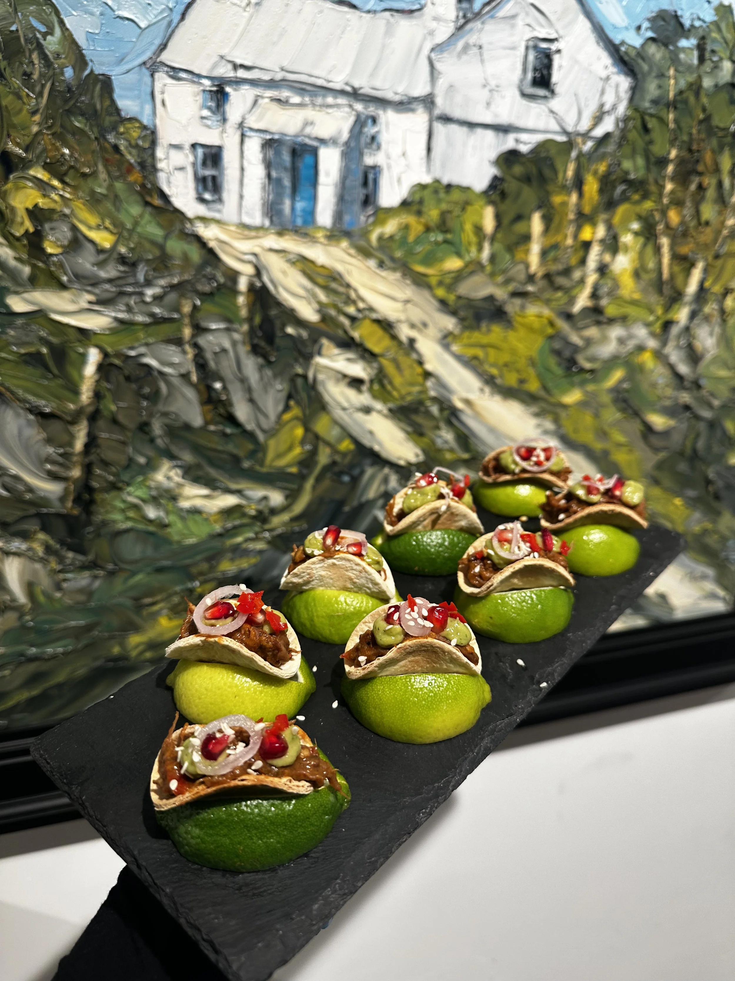 Mini tacos with meat, onions, cucumbers, and red pomegranate seeds on a black slate serving platter, with a painted landscape background.
