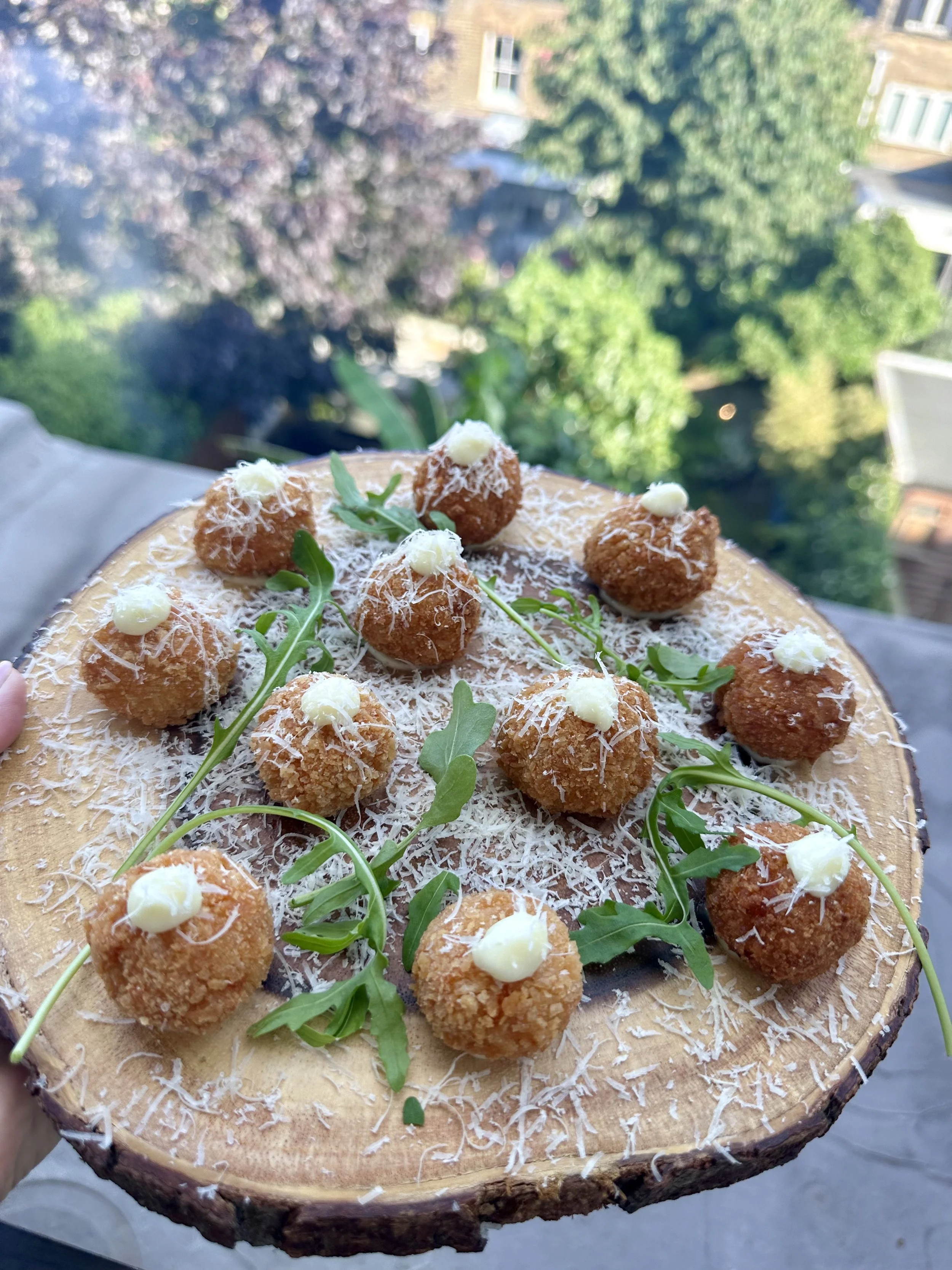 A wooden serving board with gourmet fried snacks, garnished with herbs and white sauce dots, held outdoors with trees and buildings in the background.