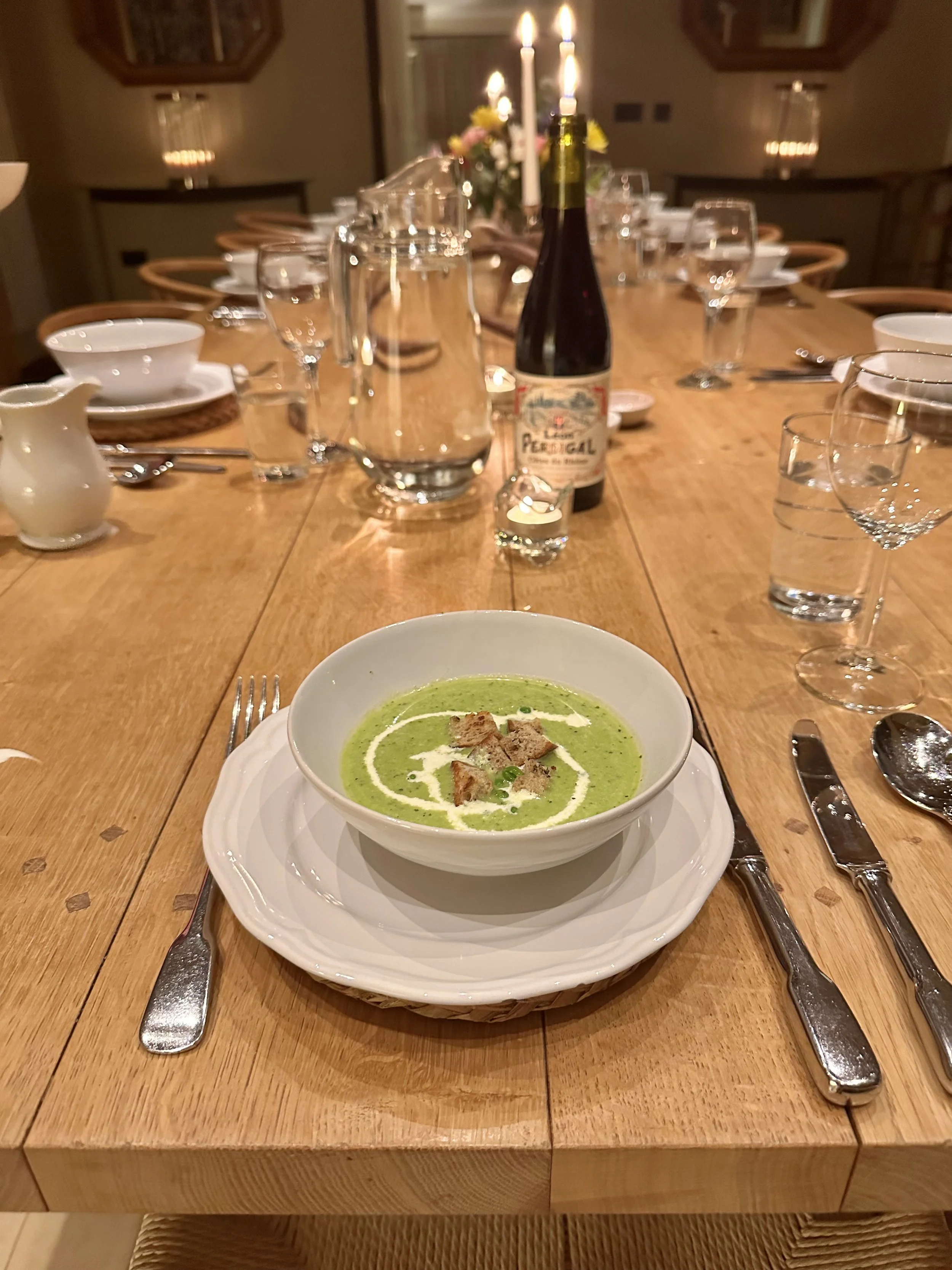 A bowl of green soup garnished with cream and croutons on a white plate, set on a wooden dining table with cutlery, glasses, a pitcher of water, a small creamer, and a bottle of wine, in a warmly lit dining room.