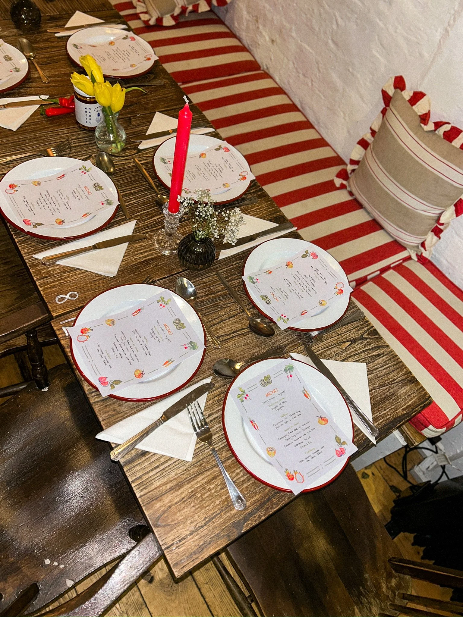 A decorated dining table set for a meal with plates, silverware, napkins, and menus, featuring yellow tulips in a small vase, a tall red candle, and small white flowers, with a red-striped cushion and pillow on a bench along a white brick wall.