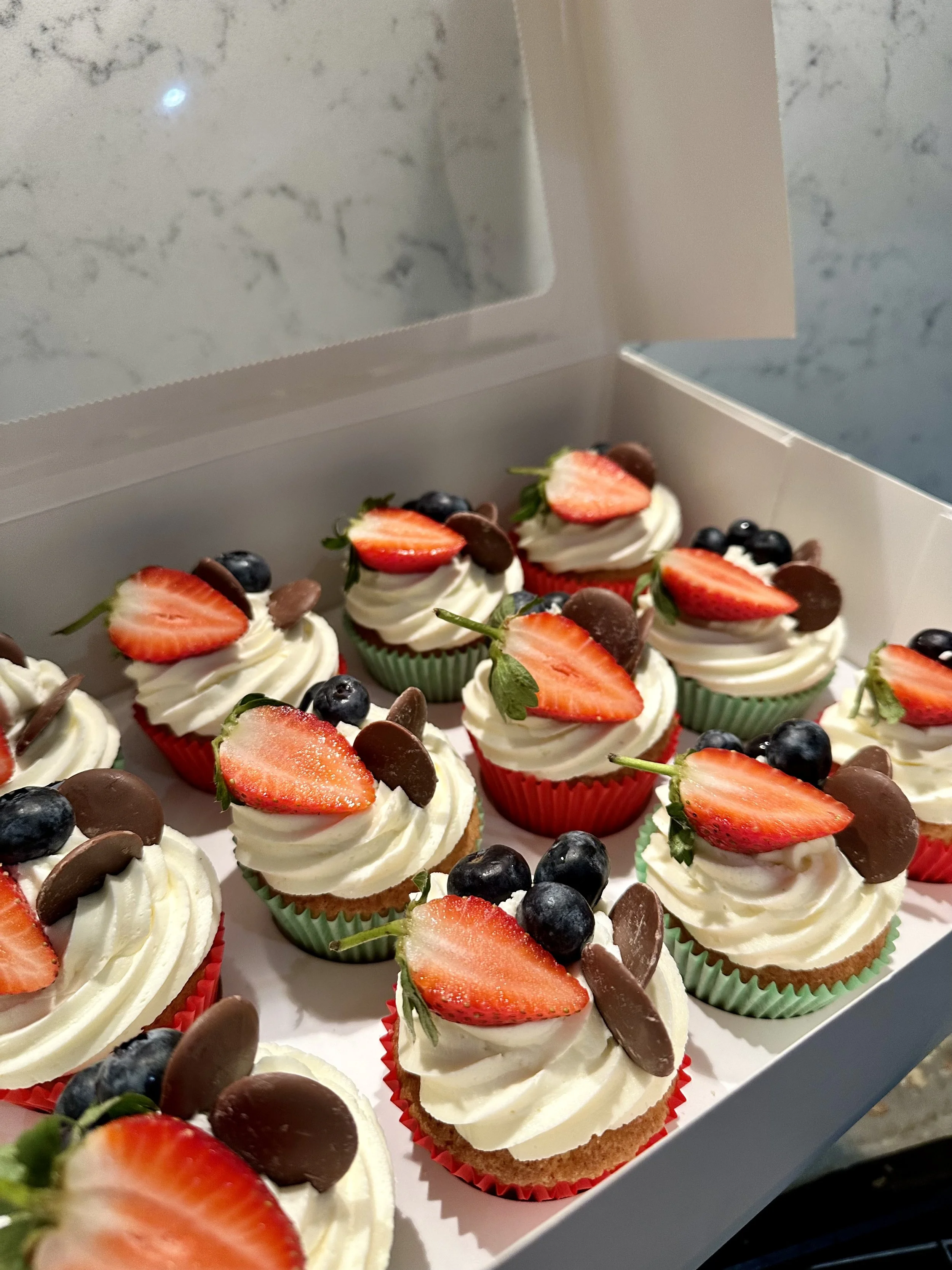 A box of cupcakes topped with white frosting, sliced strawberries, blueberries, and chocolate chips.