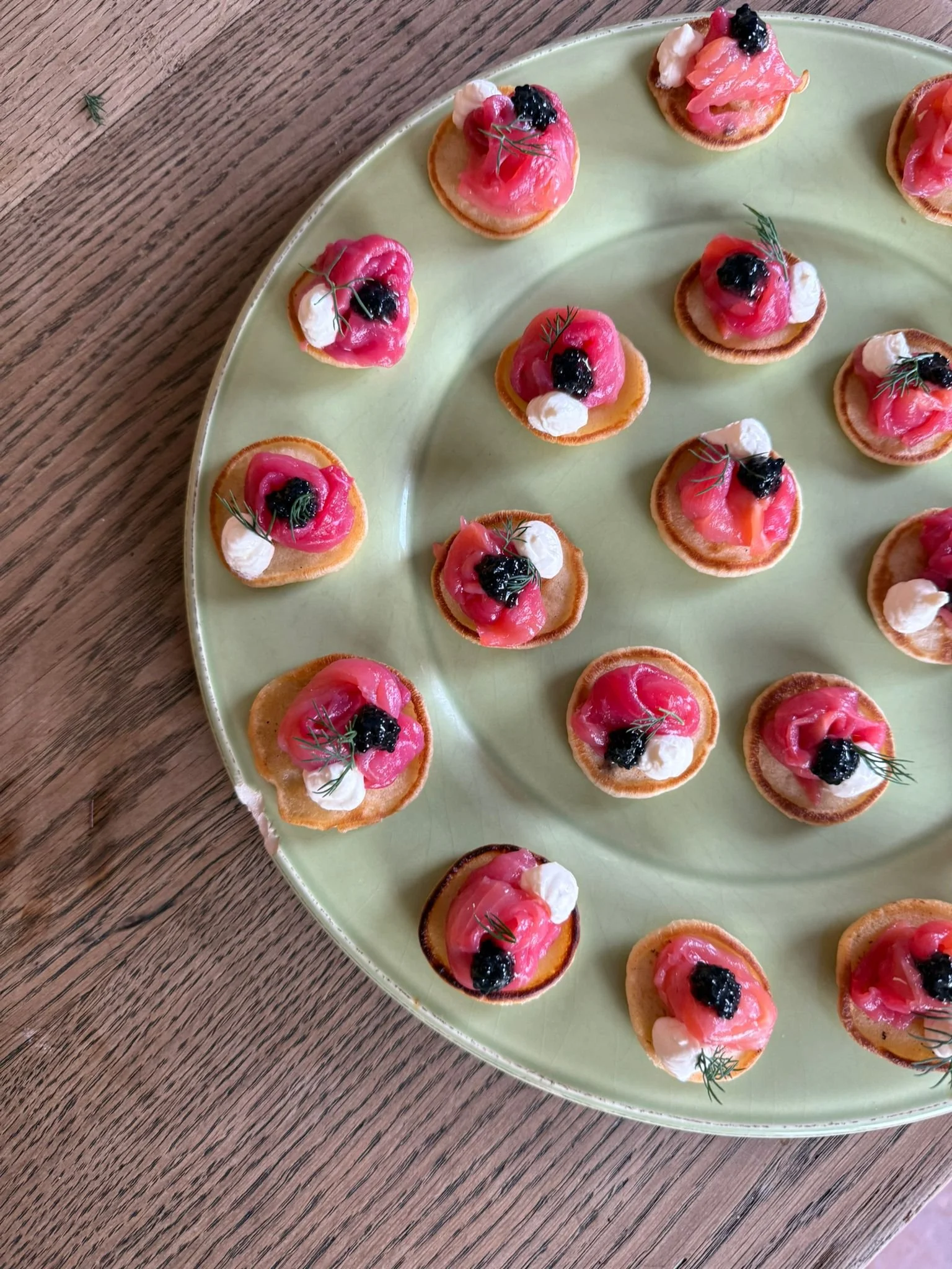Small canapé appetizers with pink smoked salmon, dollops of white cream, black caviar, and fresh dill on round crispy bases, arranged on a light green ceramic platter.
