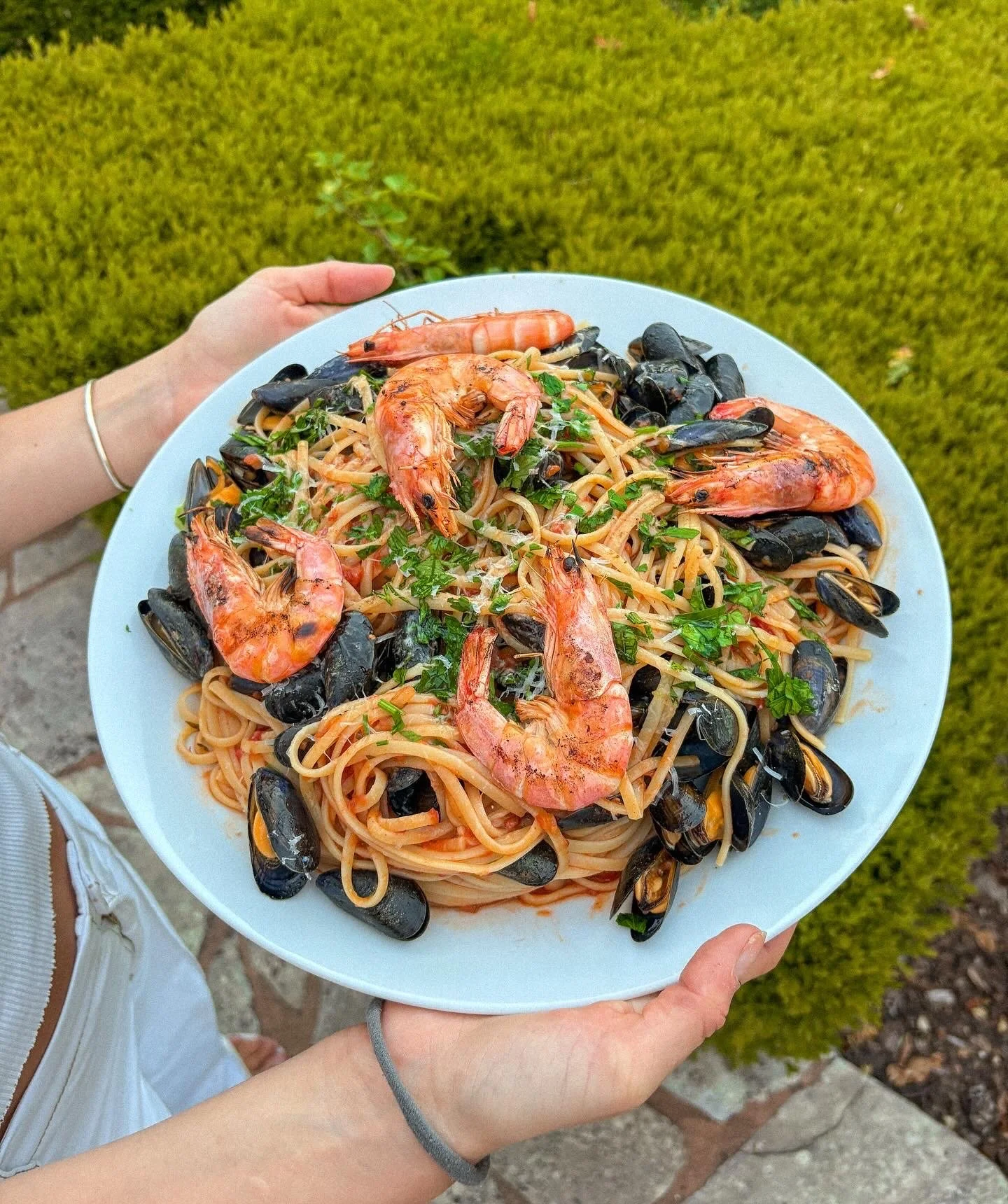 Person holding a plate of seafood pasta with shrimp and mussels outdoors on a green lawn.