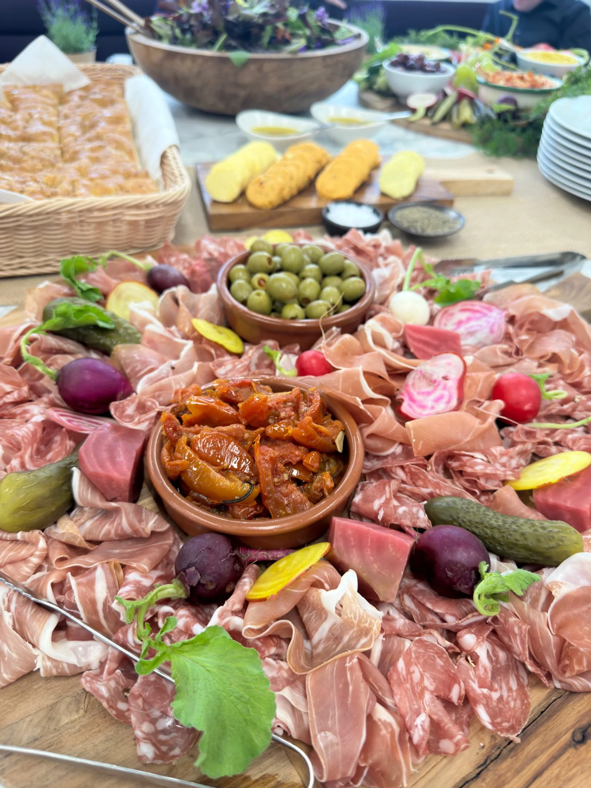 A charcuterie platter with assorted meats, cheeses, bread, and vegetables on a wooden board, surrounded by bowls of olives, roasted peppers, and other dips, with a variety of side dishes in the background.