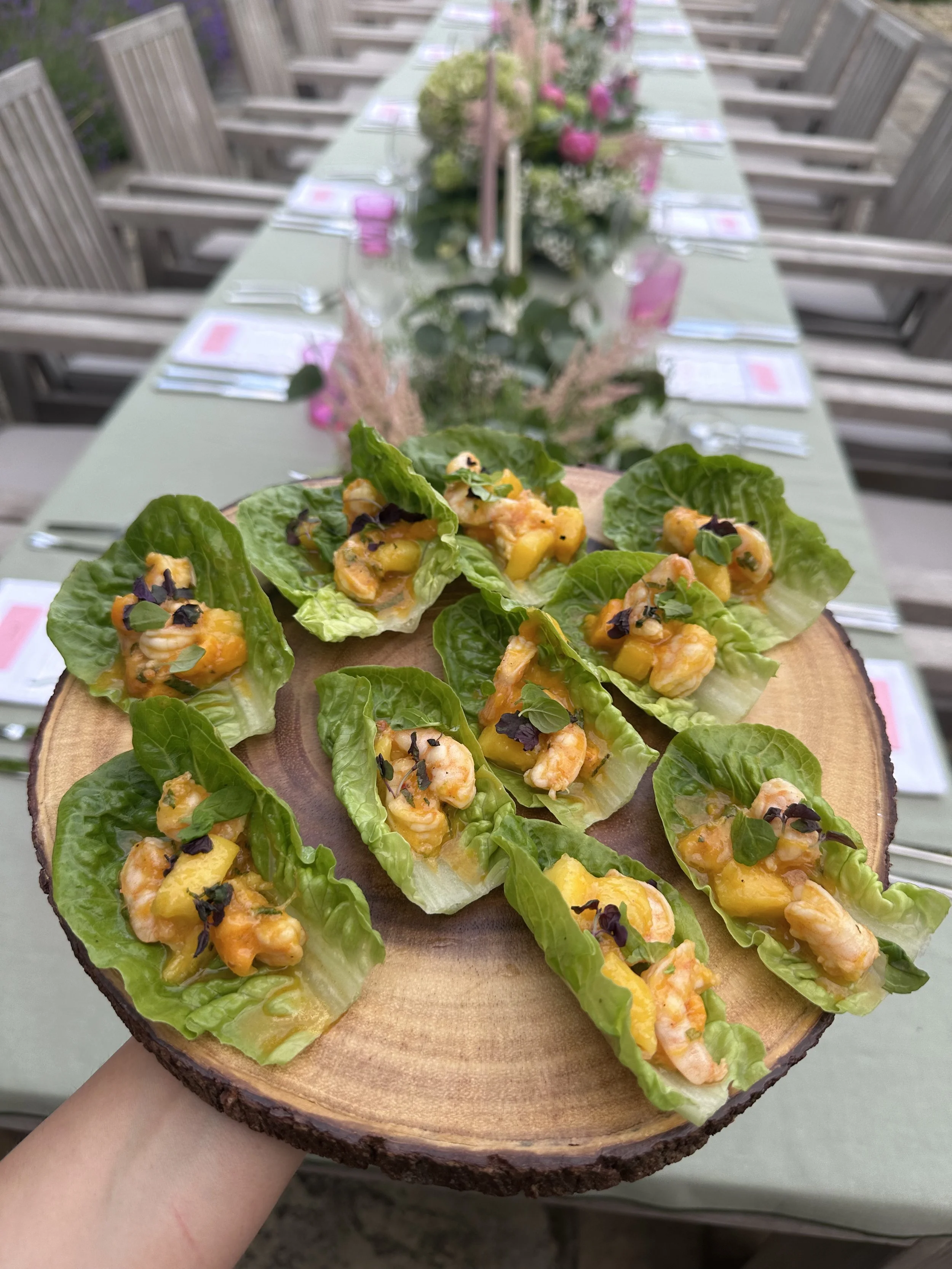 Appetizer of shrimp and mango on romaine lettuce leaves arranged on a wooden platter, with an outdoor dining table in the background.
