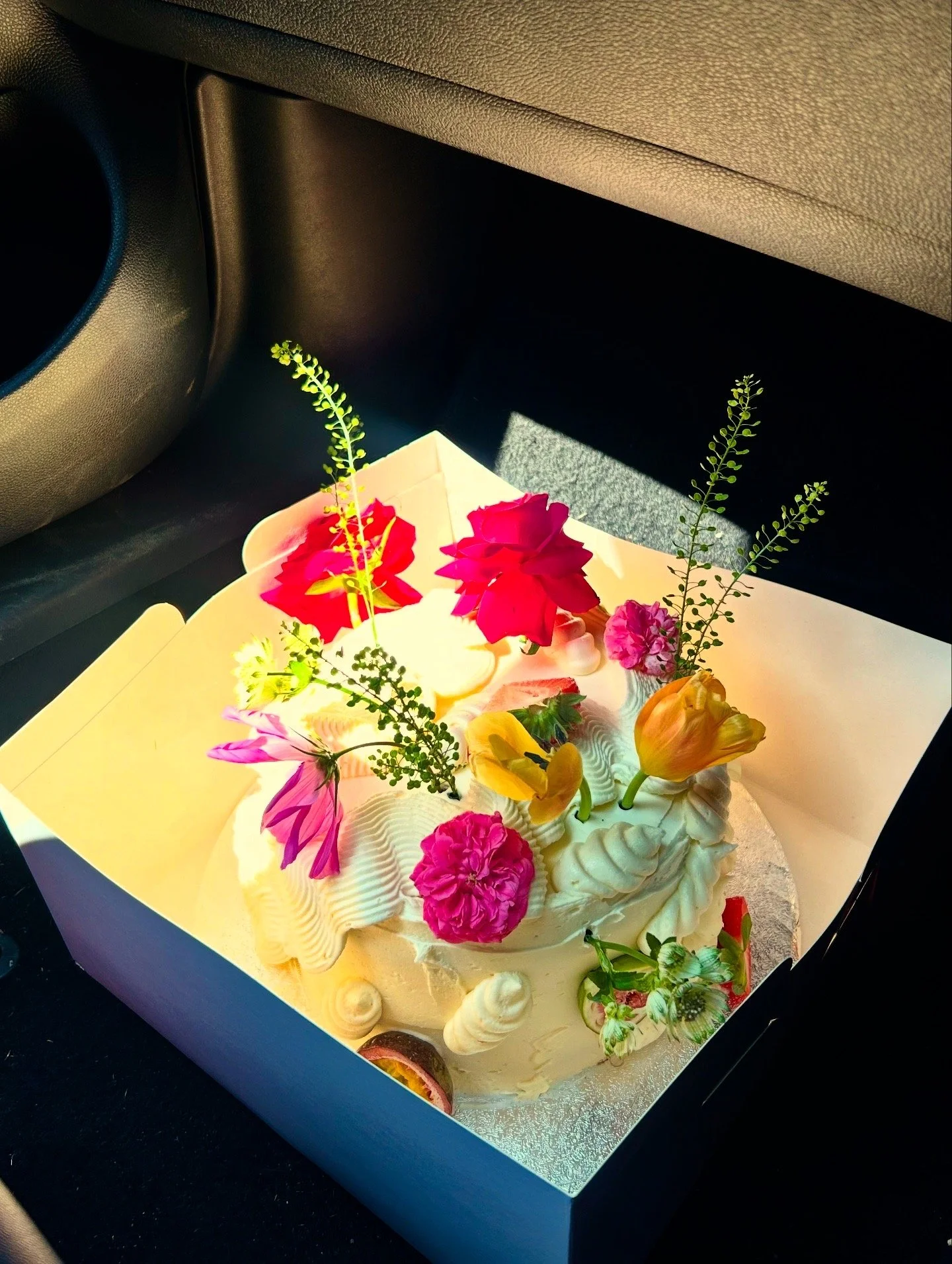 A cake decorated with colorful flowers and greenery, placed in a white box on a car seat.