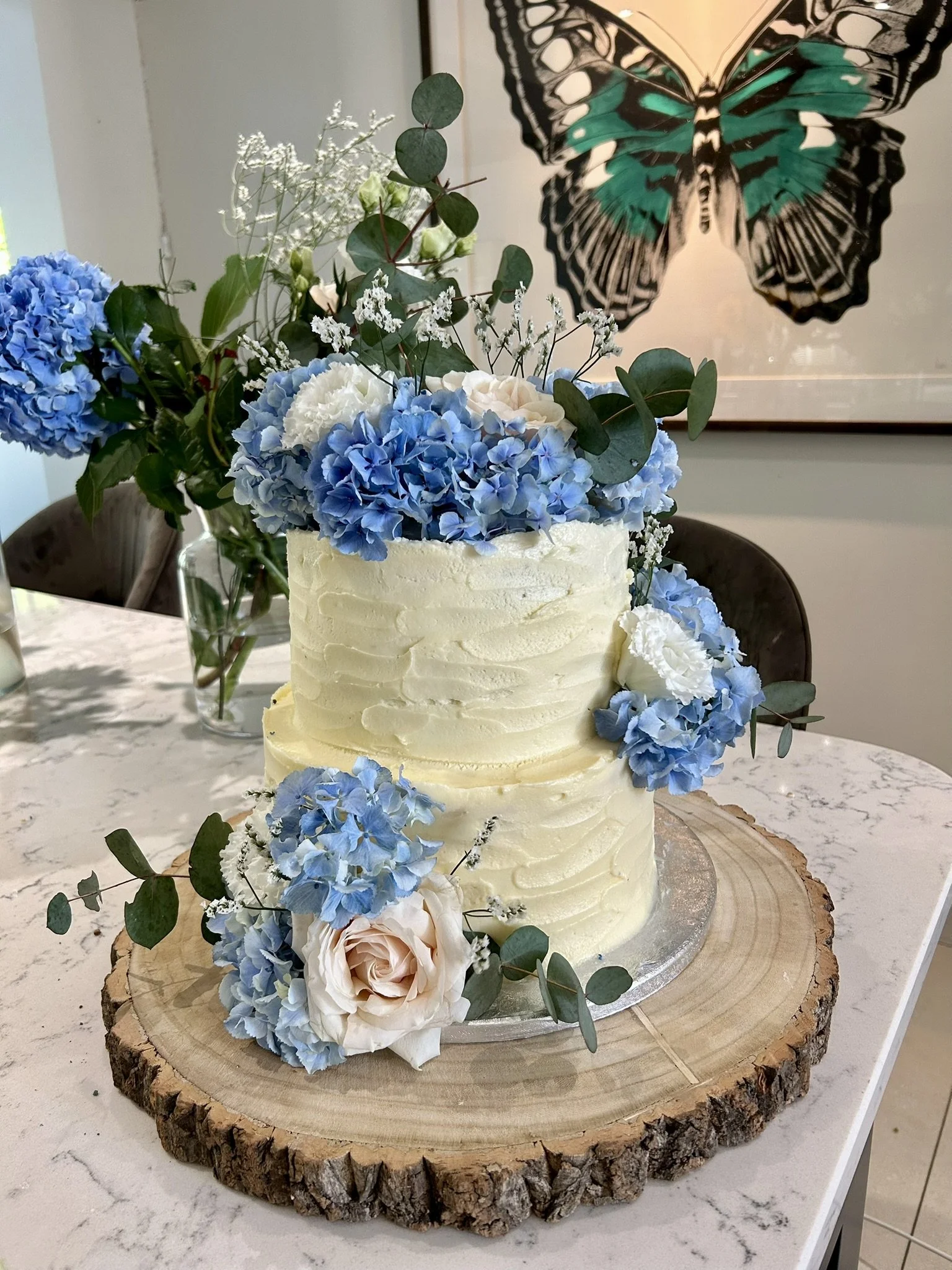 A two-tier ivory wedding cake decorated with blue hydrangeas, white roses, and greenery, placed on a wooden slab on a white marble table.