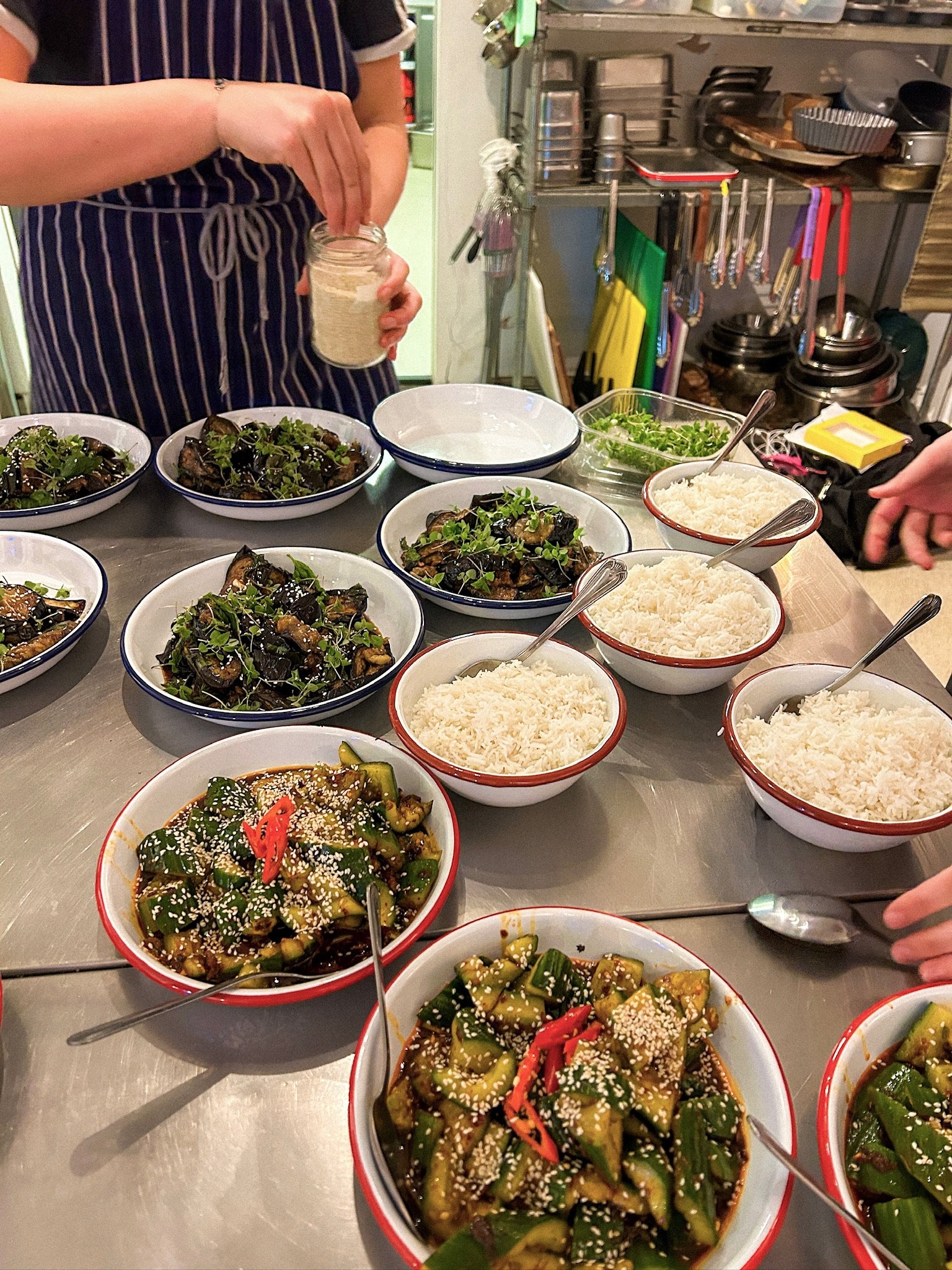 Bowls of stir-fried vegetables with sesame seeds, bowls of white rice, and bowls of cooked eggplant with garnishes on a stainless steel table in a kitchen.