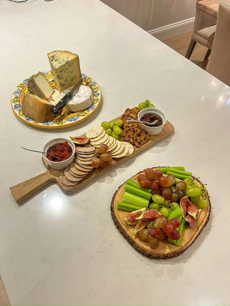 Cheese platter with various cheeses, crackers, grapes, fig slices, and jam, along with a fruit and vegetable snack board with grapes, figs, celery, and crackers.