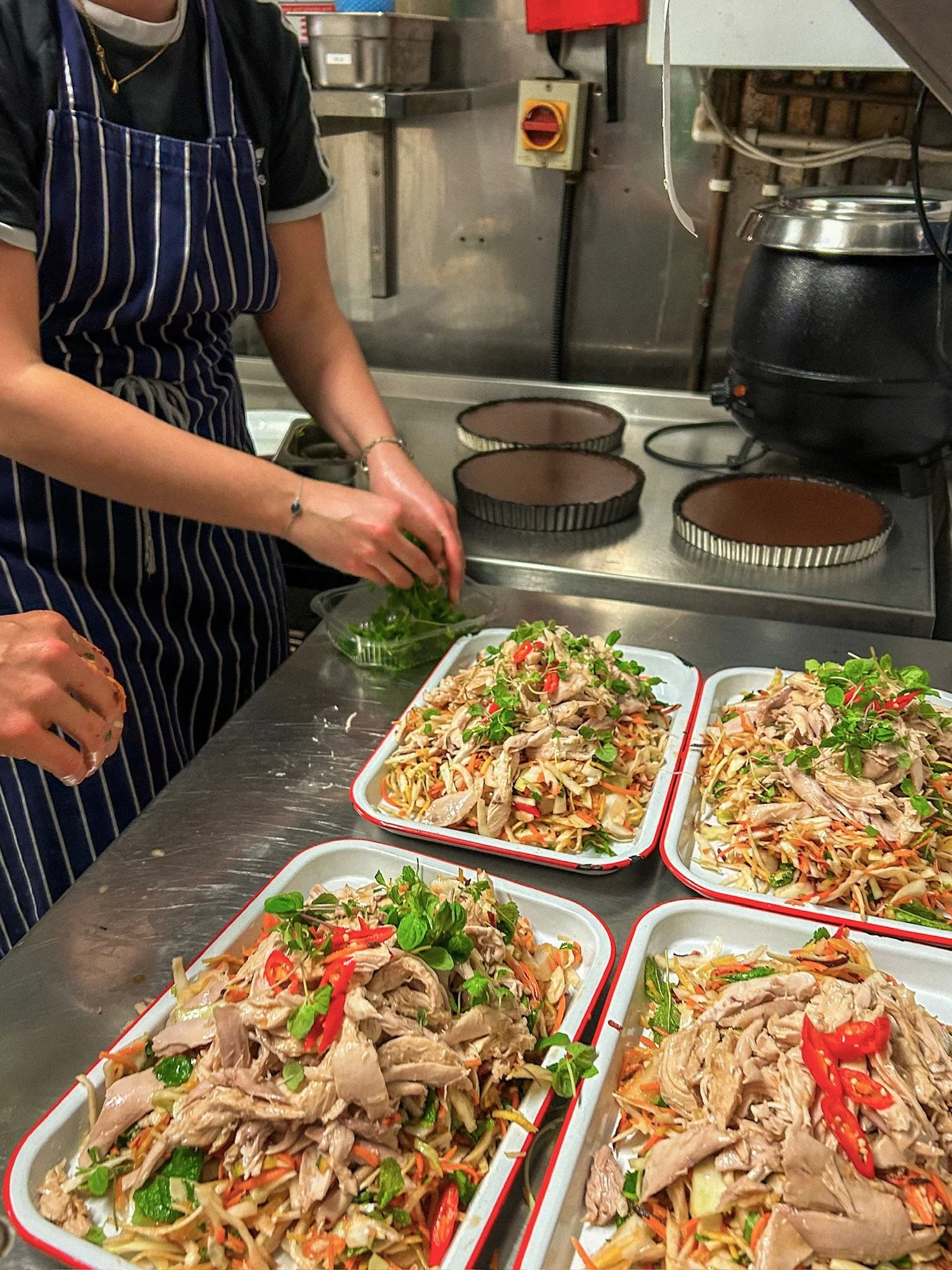 Chef garnishing platters of shredded chicken salad with herbs and sliced red peppers in a commercial kitchen.