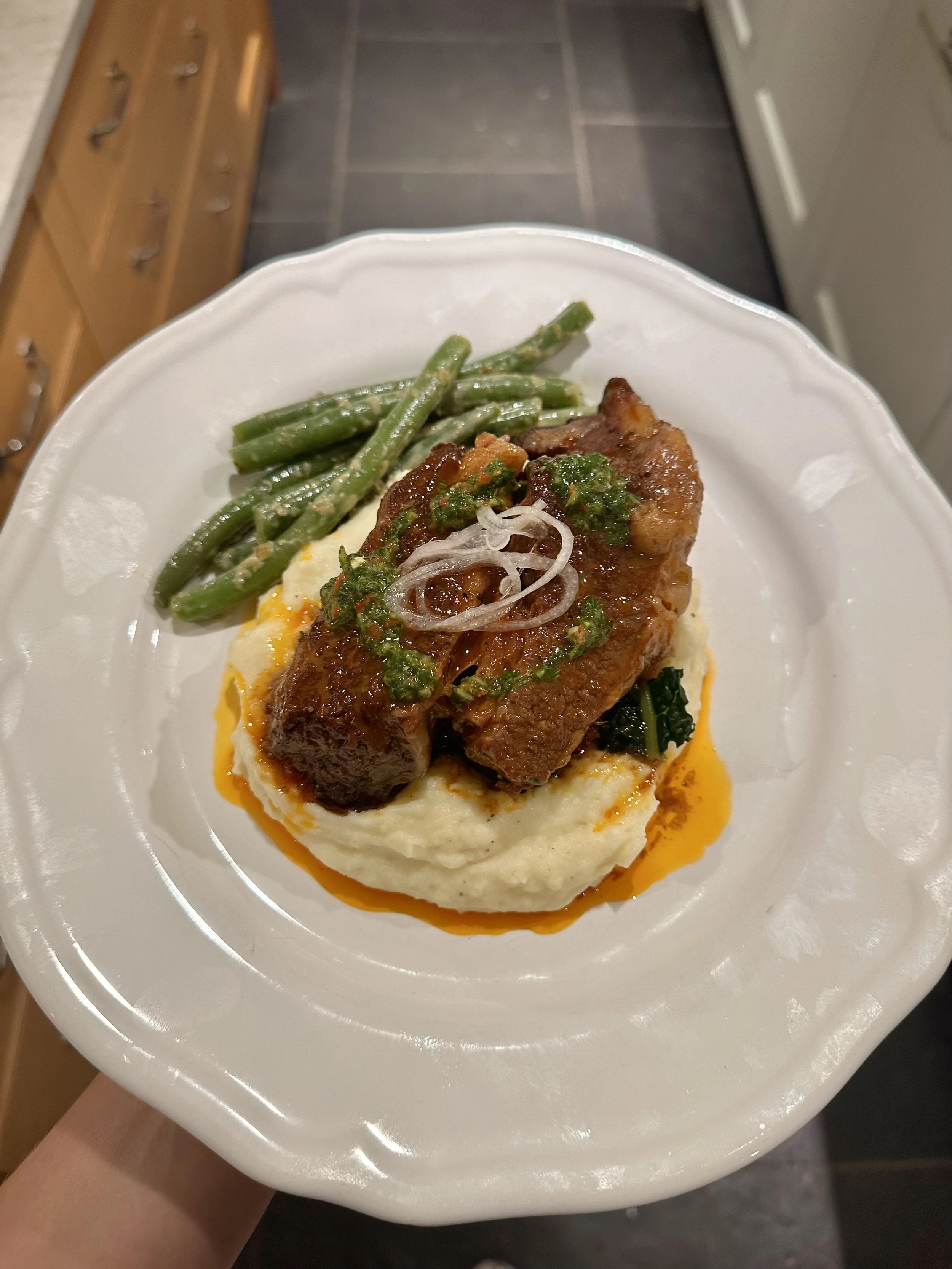 Plate of mashed potatoes topped with a cooked meat portion, green beans, and gravy with herbs and onions.