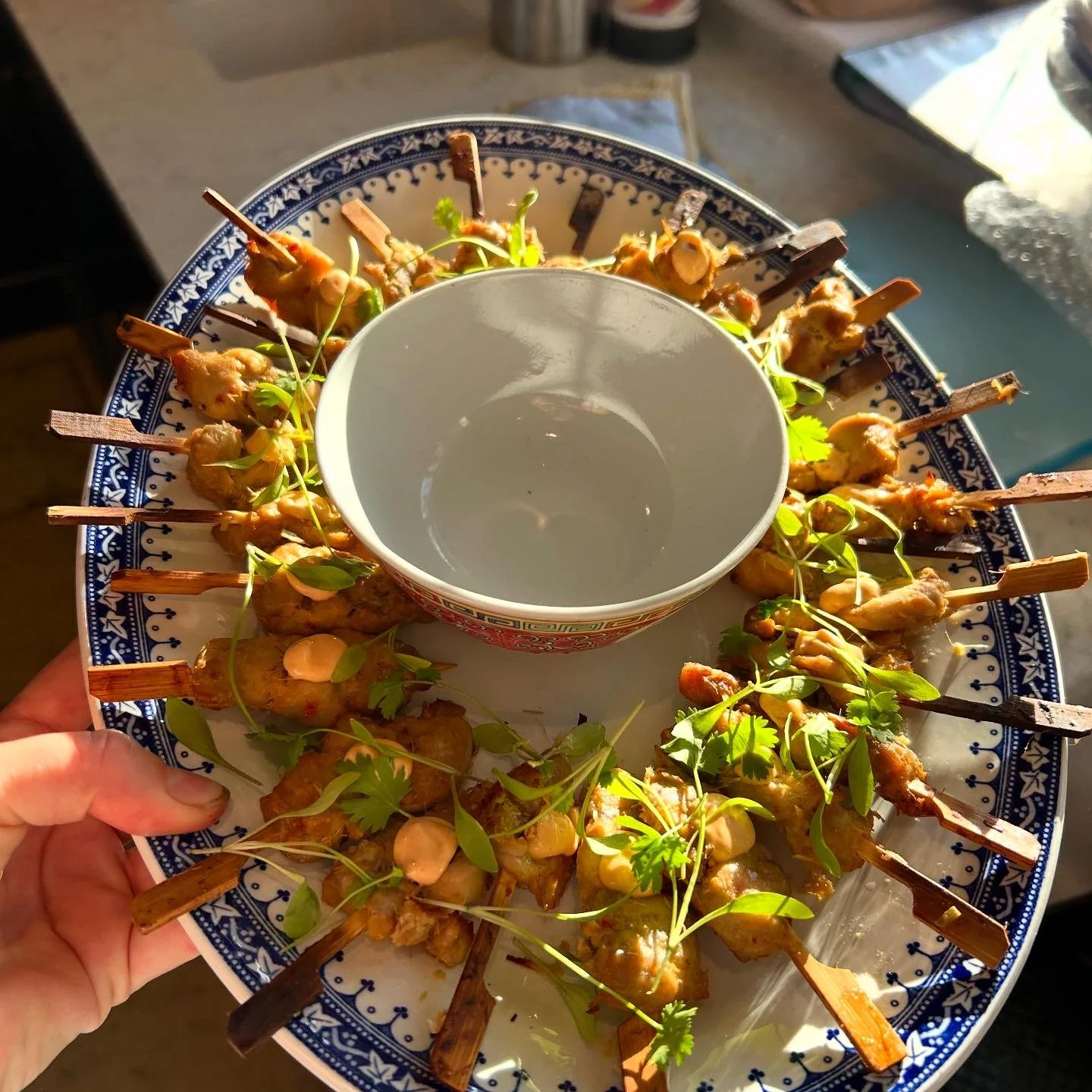 A round blue and white patterned serving plate filled with skewered grilled chicken pieces garnished with microgreens, and a white bowl in the center.