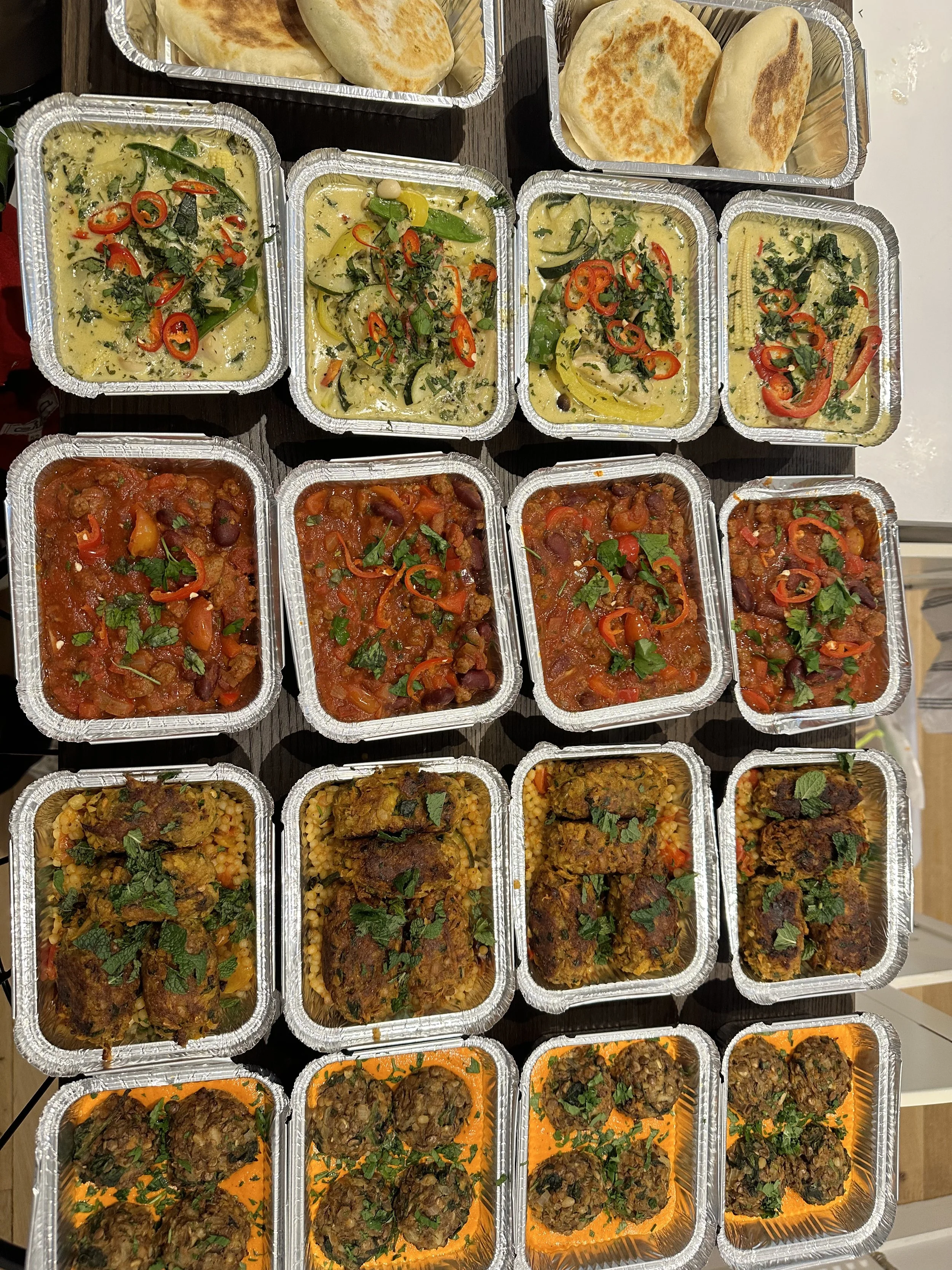 Array of Indian food dishes in foil containers, including naan bread, various curries, rice dishes, and meatballs, garnished with herbs and sliced chili peppers.