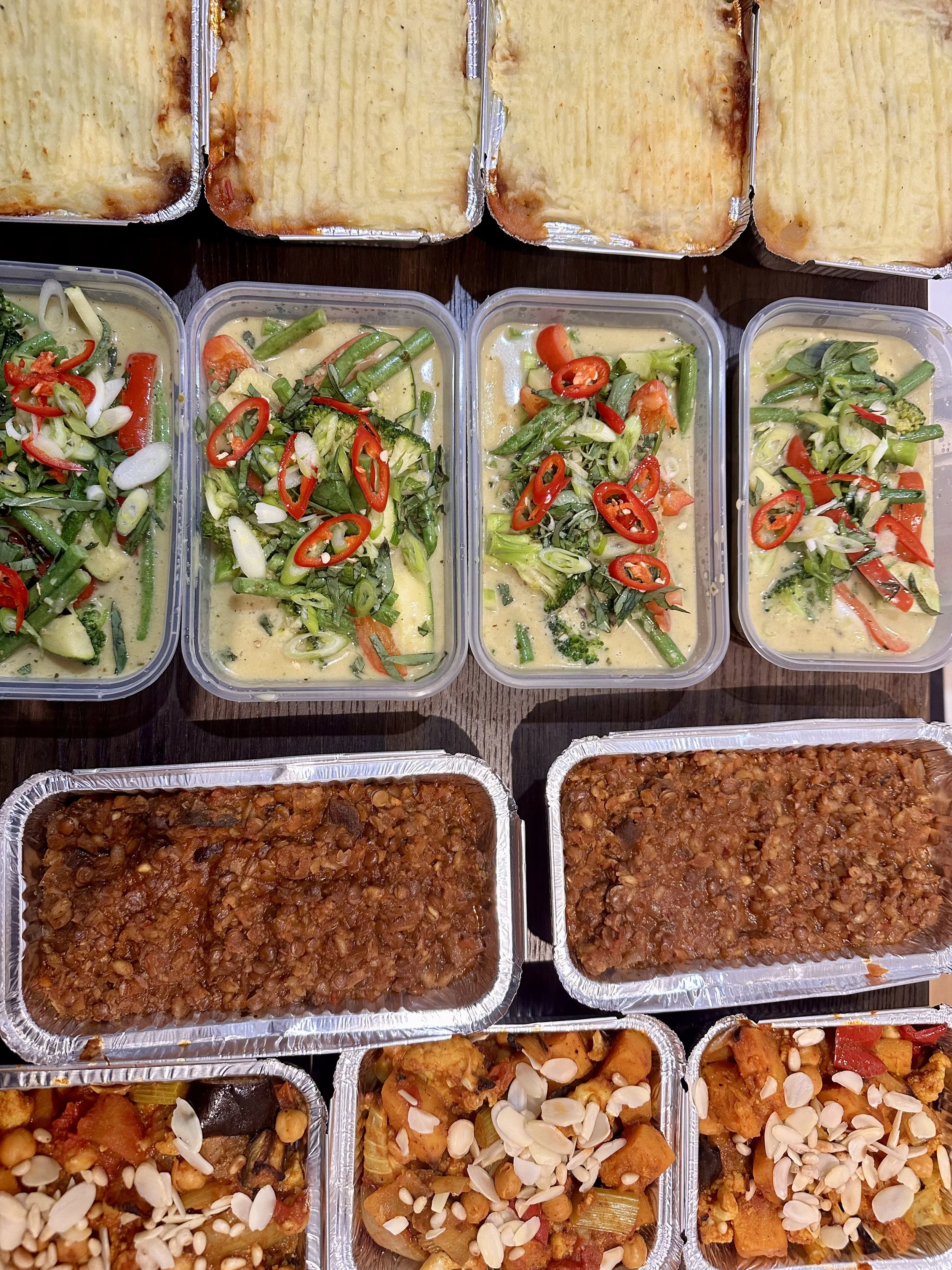 Assorted prepared foods on a table, including trays of baked casseroles, containers of vegetable curry with chopped vegetables and red chilies, and many trays of baked dishes with beef and beans topped with almonds.