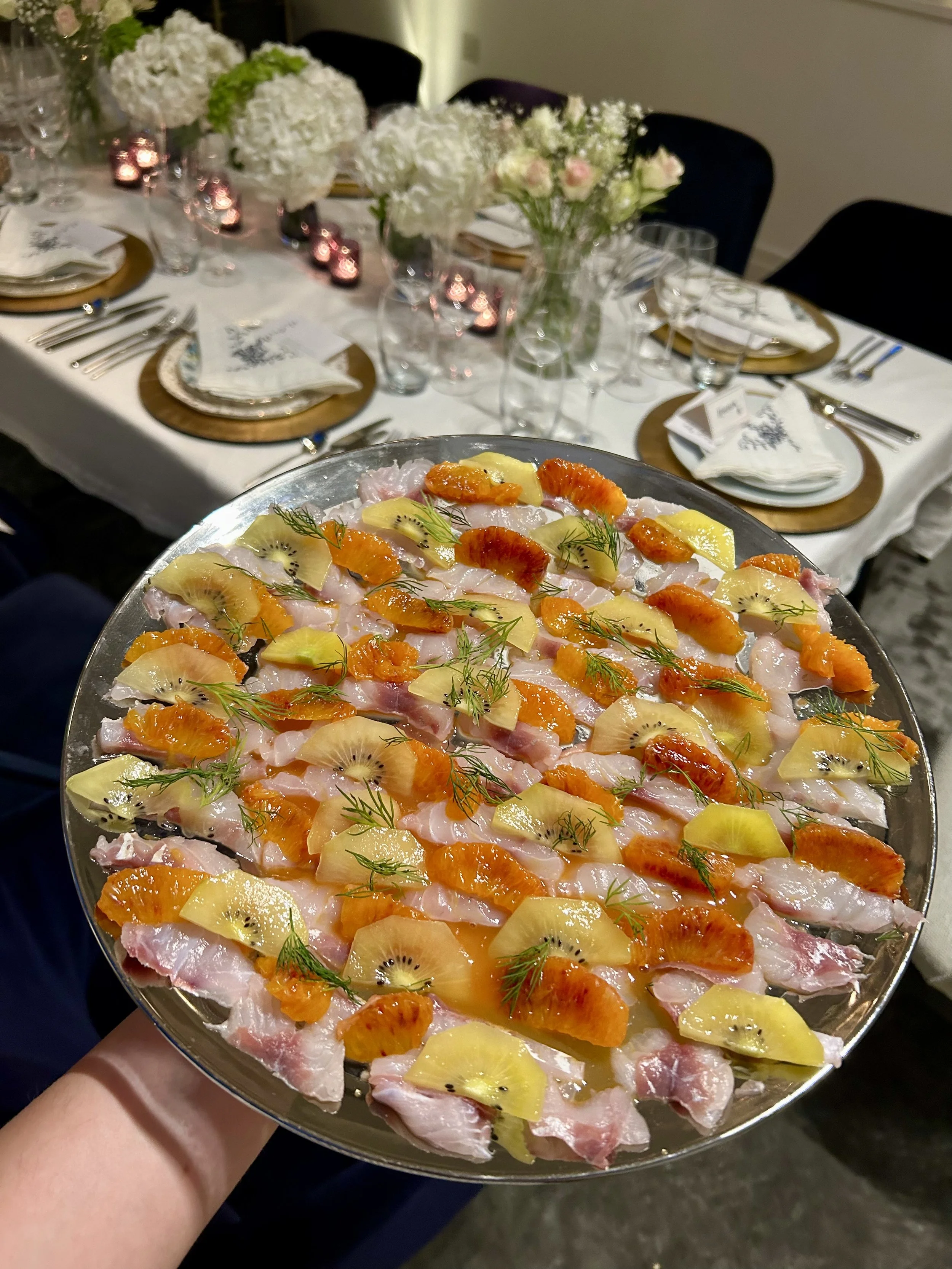 A serving platter with assorted raw fish, topped with slices of kiwi, mandarin oranges, and sprigs of dill, held in front of a decorated dining table with floral centerpieces and elegant place settings.