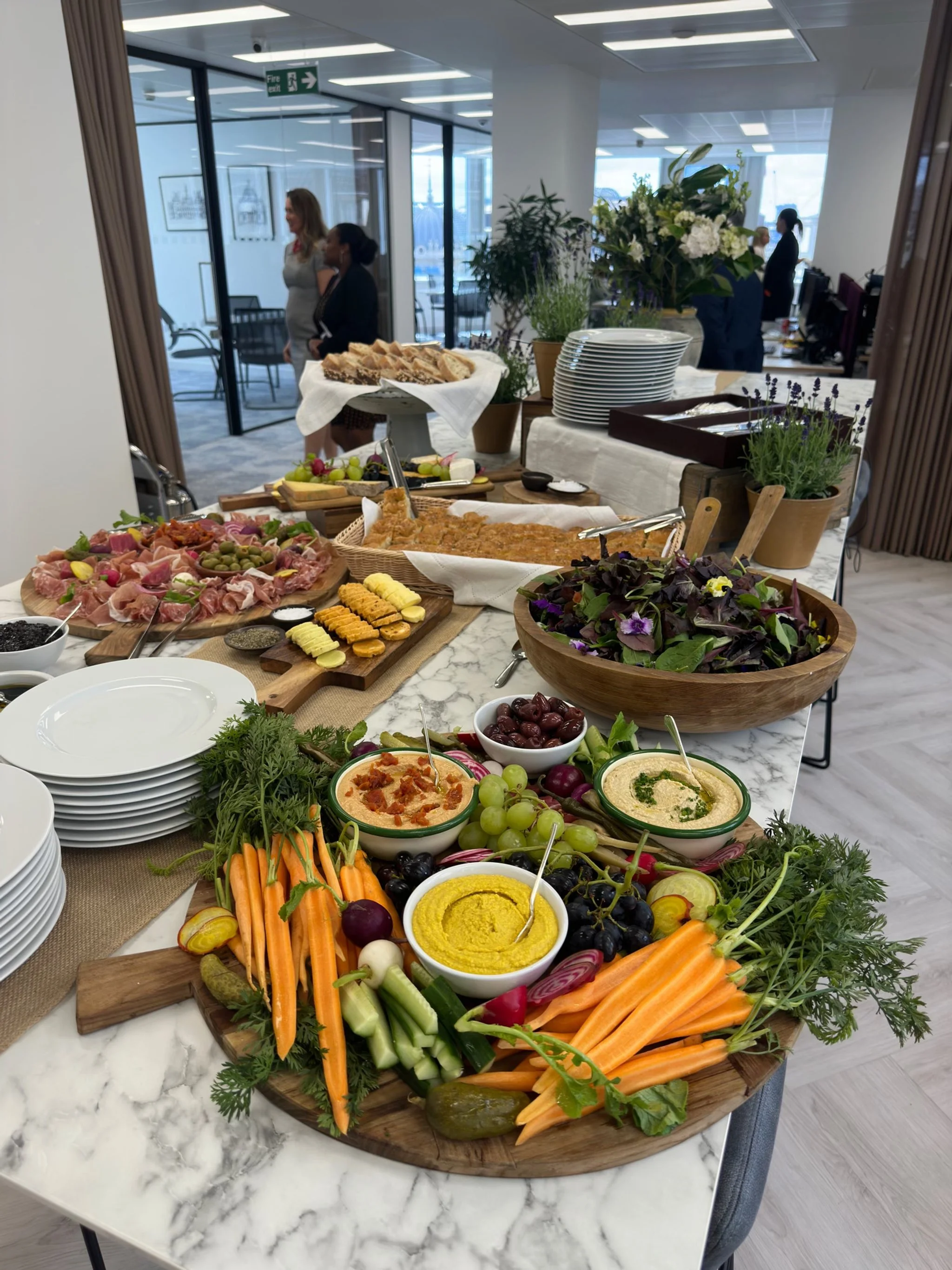 An assortment of appetizers and vegetables, including dips, grapes, carrots, and cucumbers, arranged on a white marble table at a buffet or gathering.