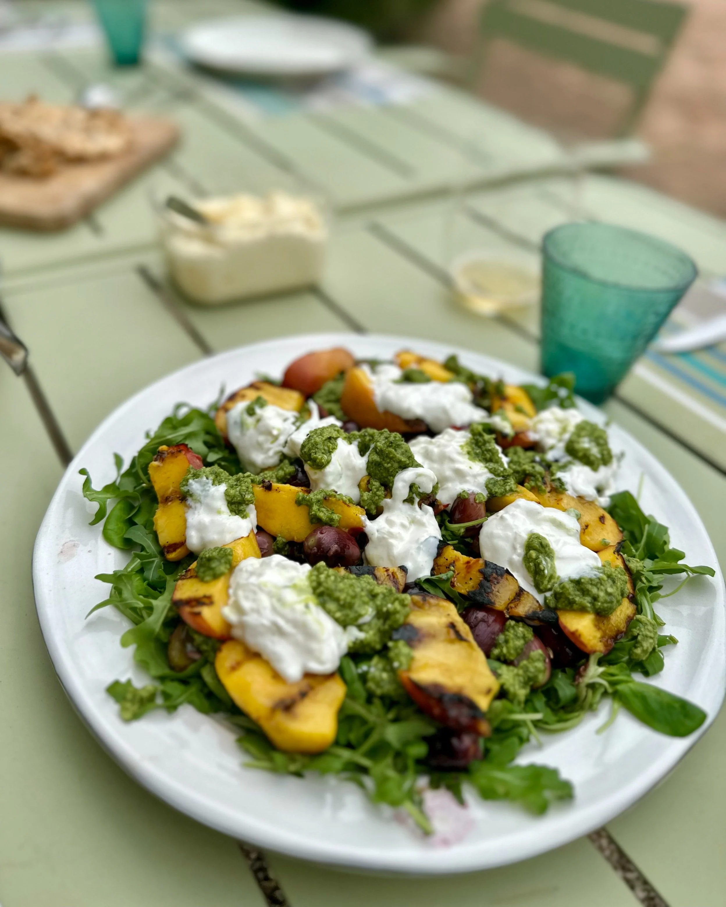 A plate of grilled peach salad with greens, white cheese, and green dressing on a light green table outdoors.