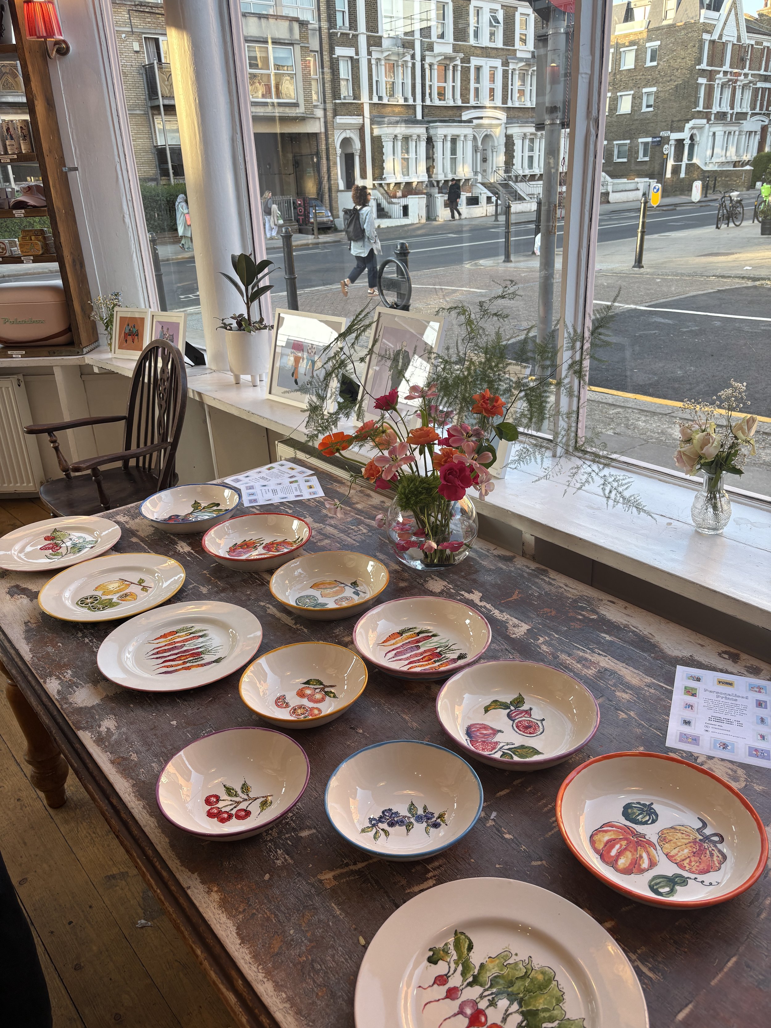 Colorful ceramic plates with vegetable and fruit illustrations are arranged on a rustic wooden table inside a shop near a large window. Flowers and framed artwork decorate the windowsill.