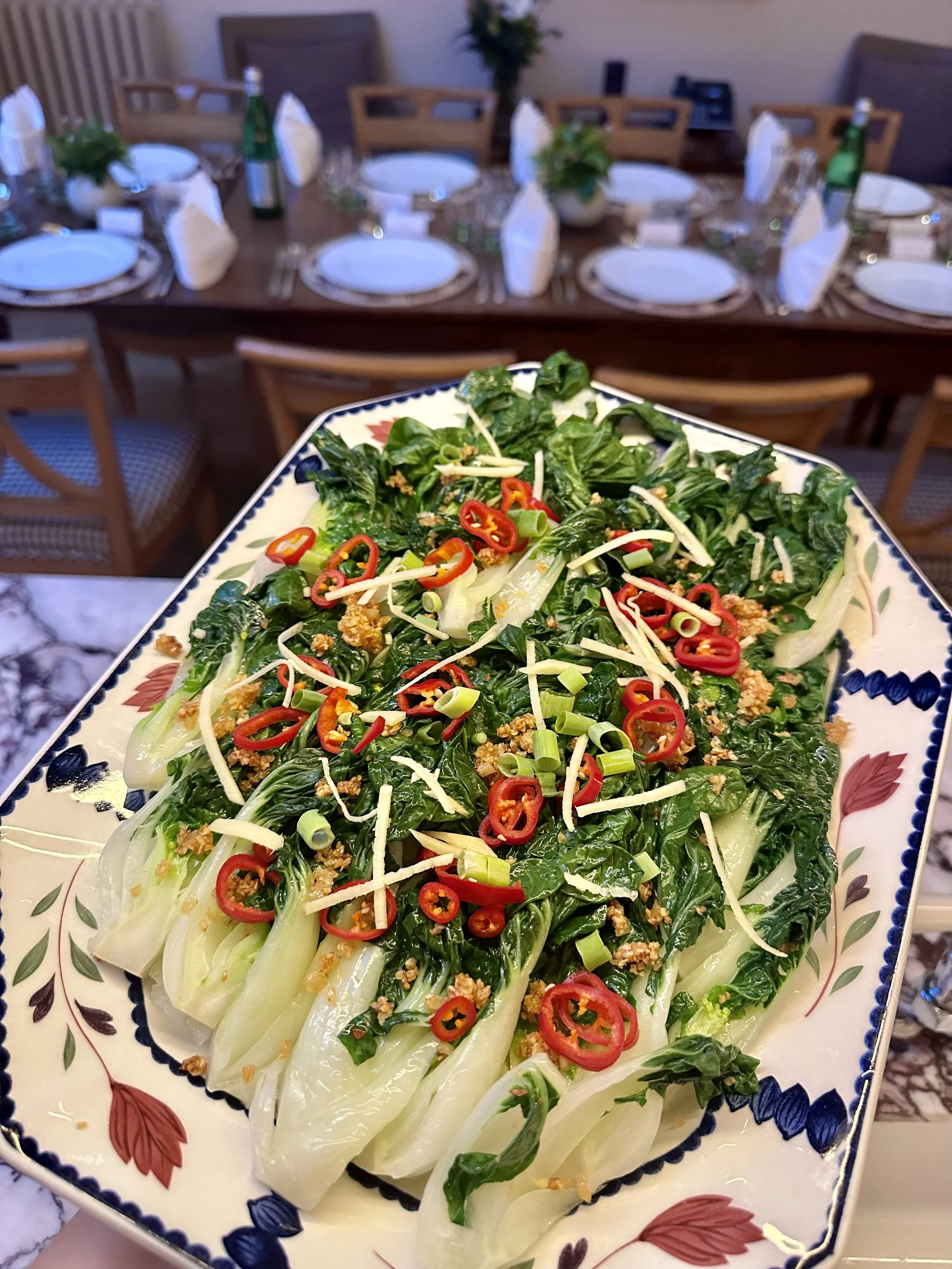 A decorative serving platter with sautéed leafy greens, topped with sliced red chili peppers, shredded cheese, chopped green onions, and crunchy garlic bits.
