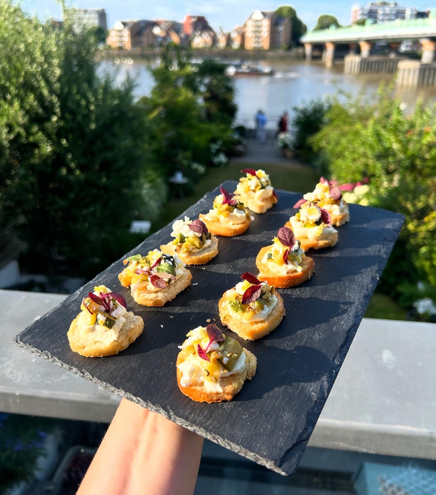A hand holding a black slate platter with twelve small appetizers topped with edible flowers against a blurred background of trees, water, and buildings.