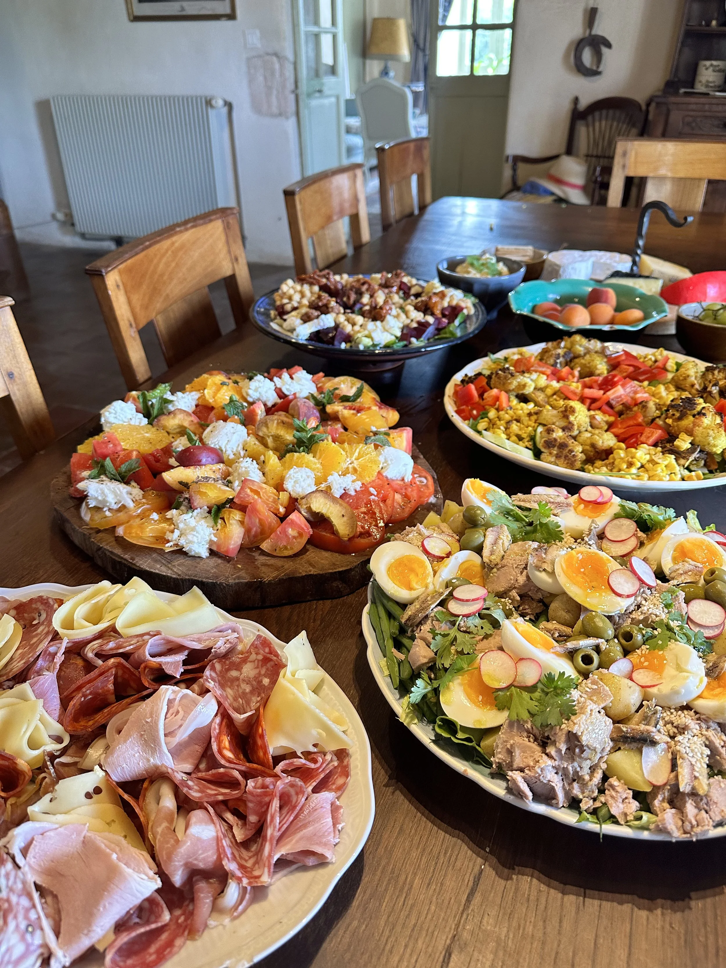 A table set with various colorful salads and cold cuts, including a tomato salad with feta cheese, a plate with halved soft-boiled eggs, a mixed salad with roasted vegetables, and an antipasto platter with meats and cheeses. In the background, there 
