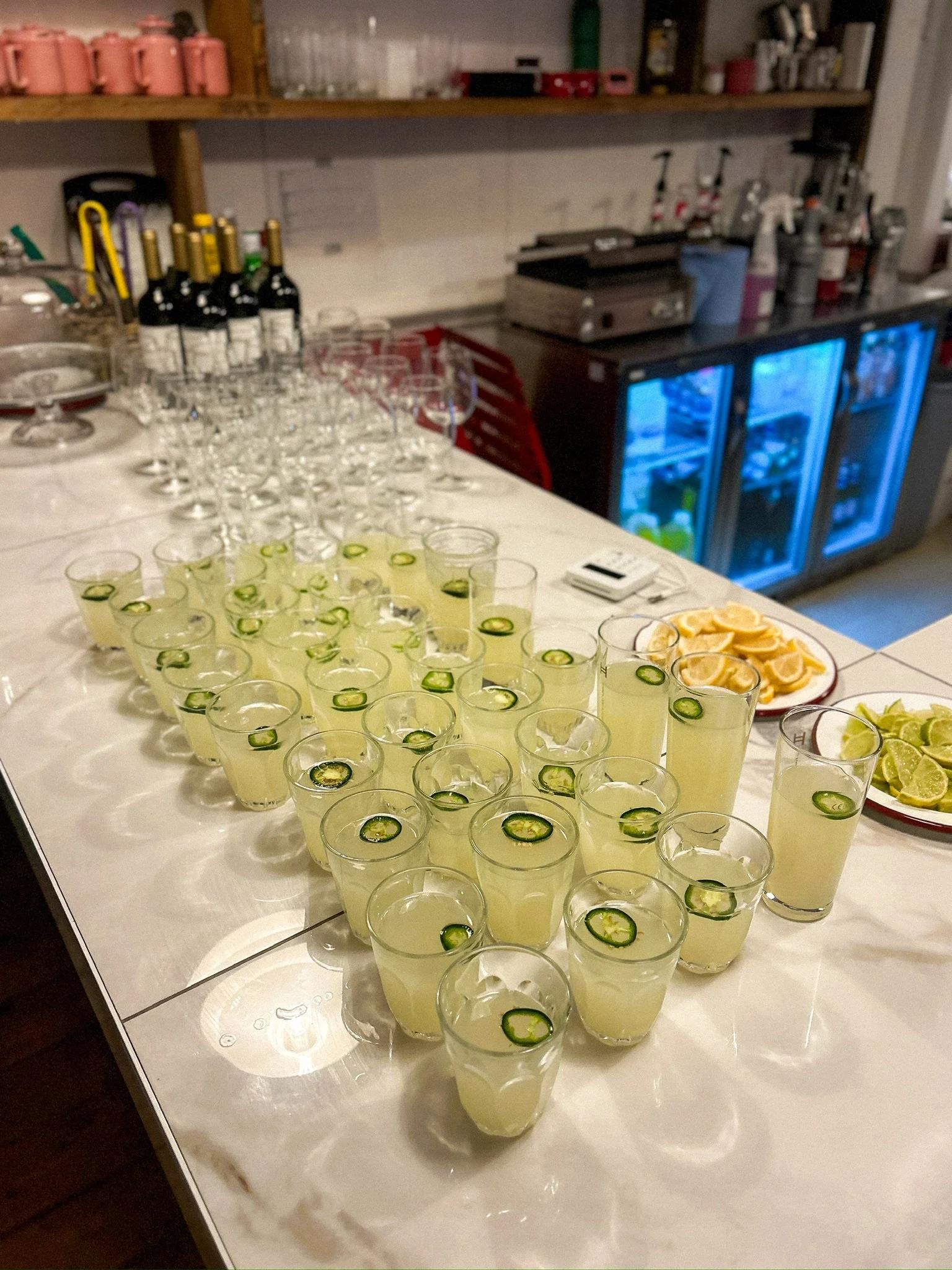 A table with rows of glasses filled with a pale yellow drink, garnished with slices of jalapeño peppers. There are plates of lemon and lime wedges nearby. In the background, bottles of wine and various bar equipment are visible.
