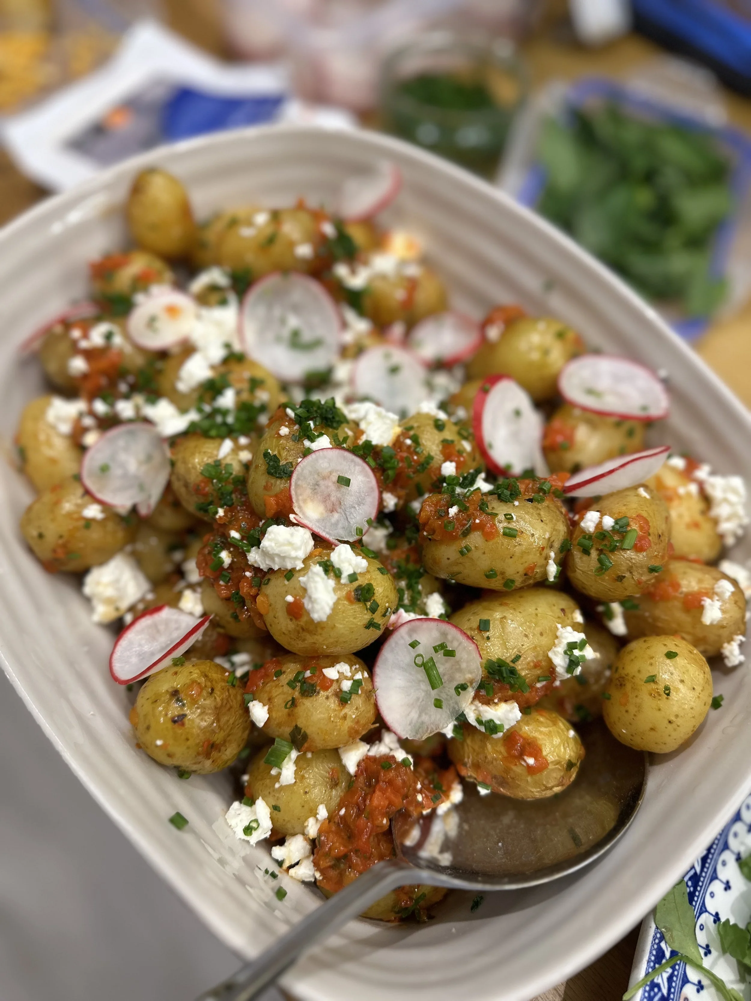 A dish of seasoned new potatoes garnished with radish slices, chopped chives, crumbled cheese, and a red pepper mixture in a white serving bowl.