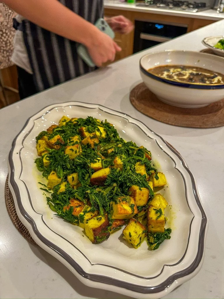 Dish of roasted sweet potatoes mixed with sautéed greens on a white platter with a black decorative border, set on a table with a bowl of soup and a green lime wedge in the background.