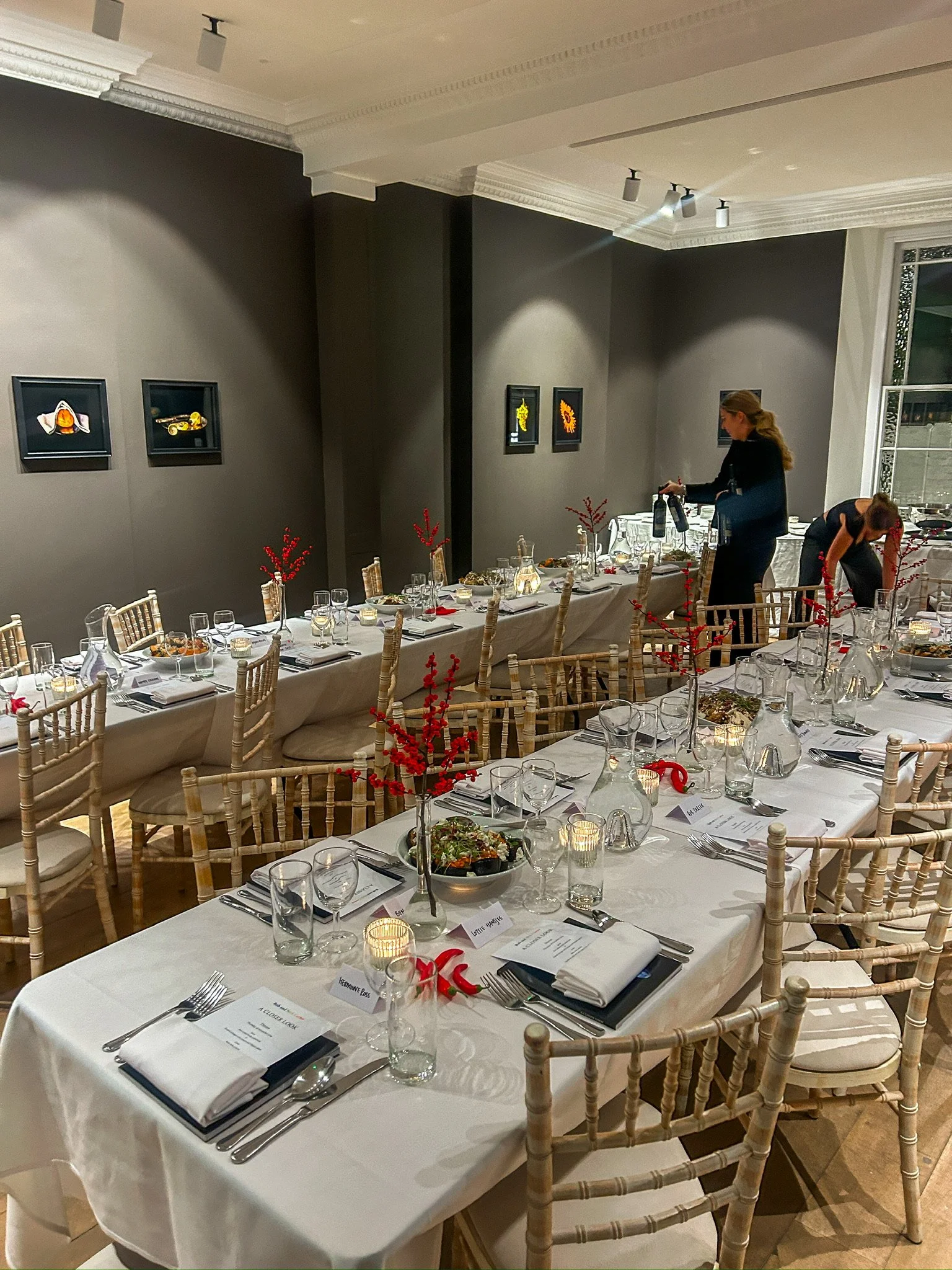 A banquet hall decorated for an event with long tables set with white tablecloths, napkins, glassware, and silverware, with red floral centerpieces and candles, and framed artwork on the walls.