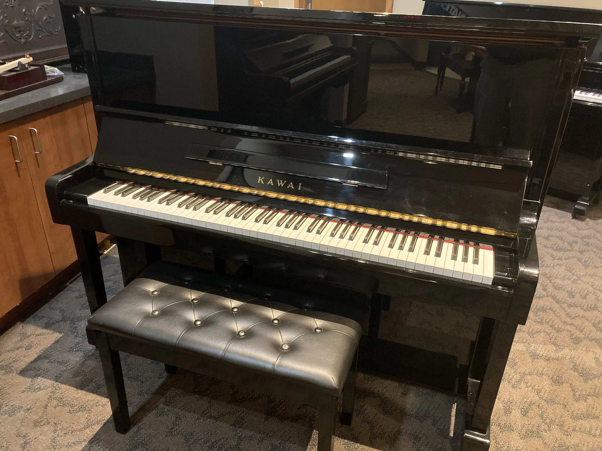 Black upright Kawai piano with a black leather bench, located in a room with carpeted flooring.