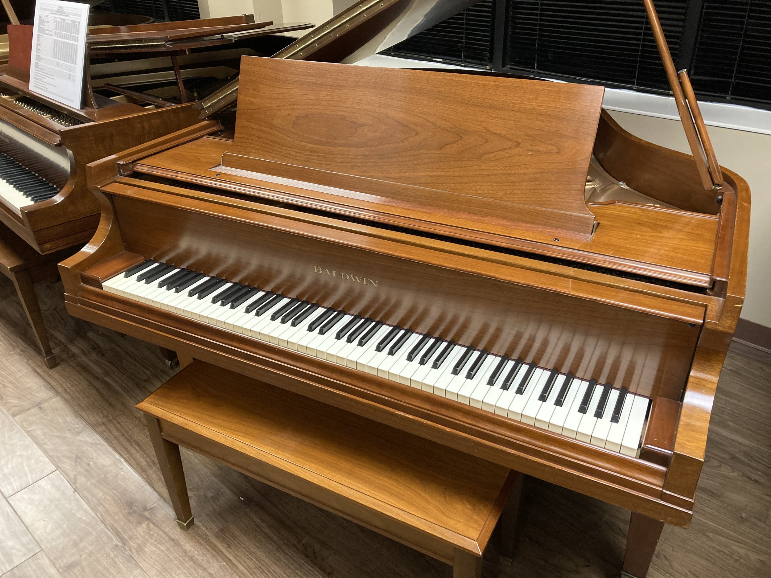 Brown Baldwin grand piano in a showroom with a partially open lid and a music sheet nearby.