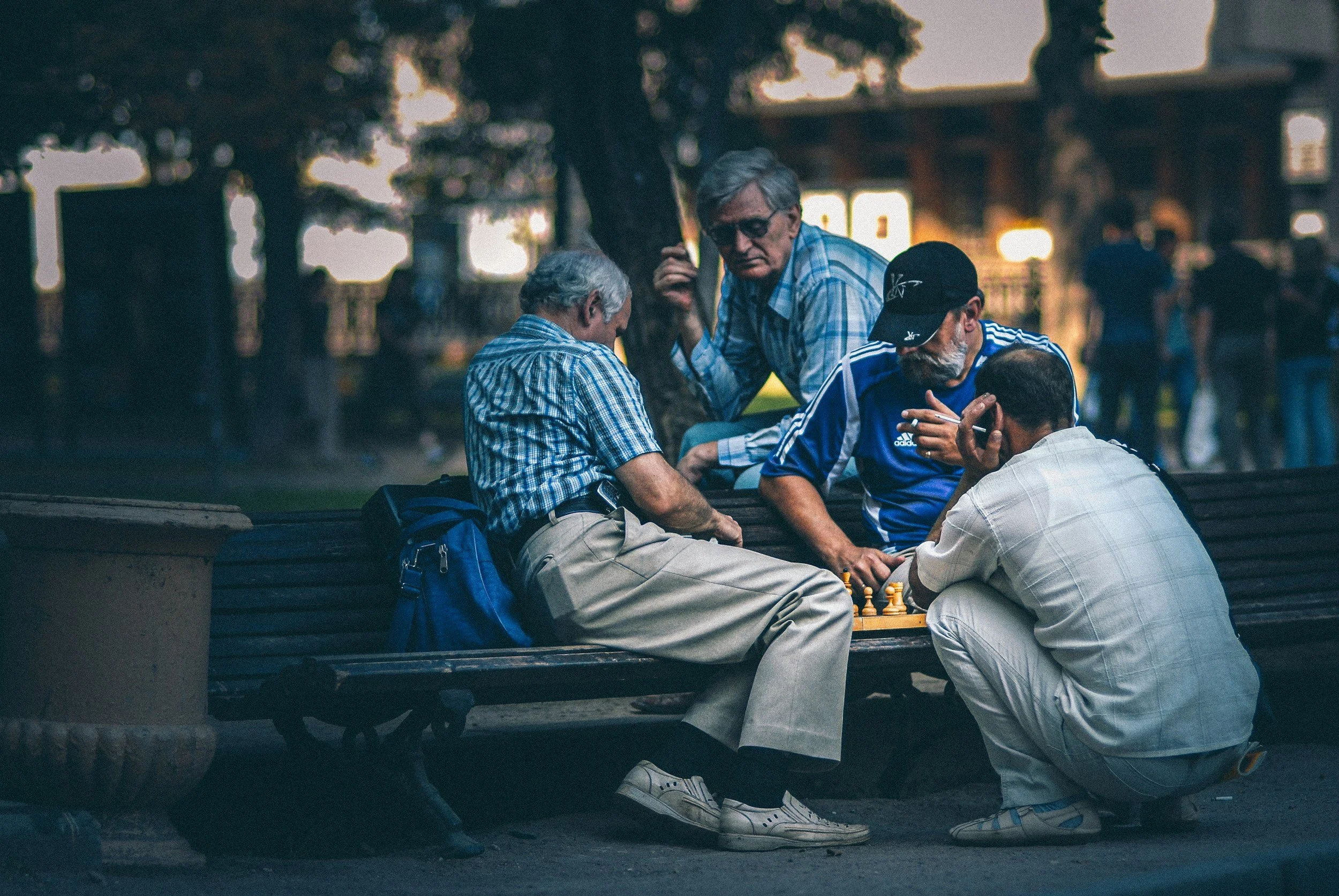 Four elderly men are playing chess on a park bench during sunset, with trees and some blurred figures in the background.