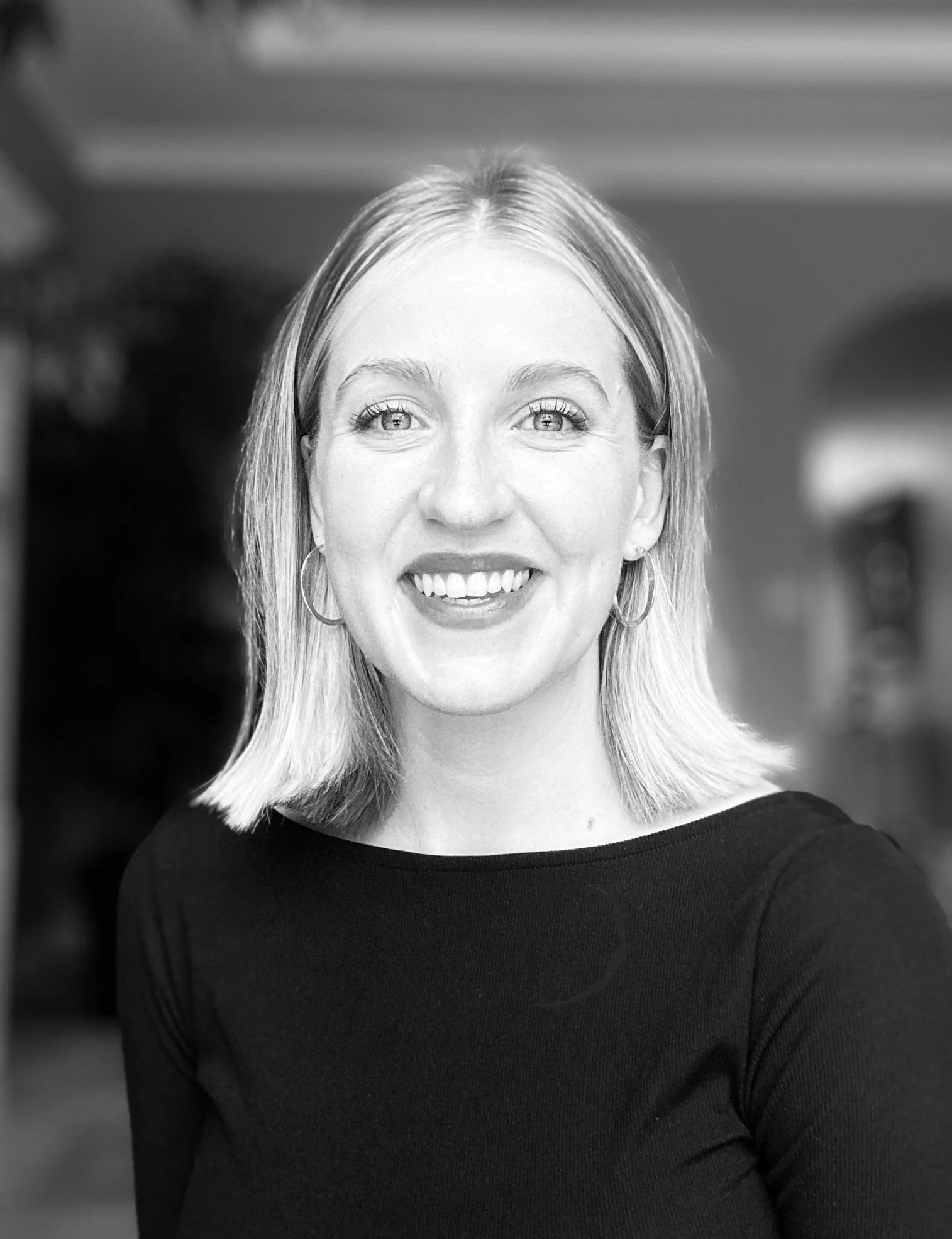 A smiling woman with shoulder-length blonde hair, wearing hoop earrings and a black top, in an indoor setting.