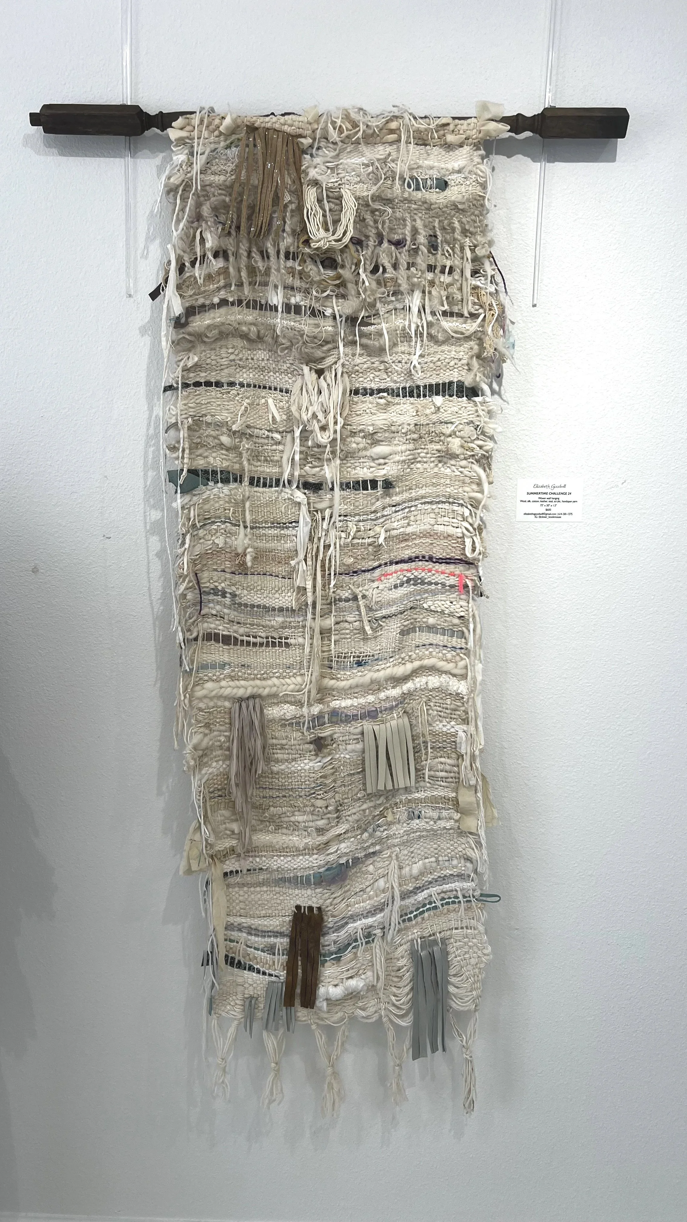 A textile art piece made of various woven and knitted fabric strips and yarns hanging vertically from a wooden rod on a wall.