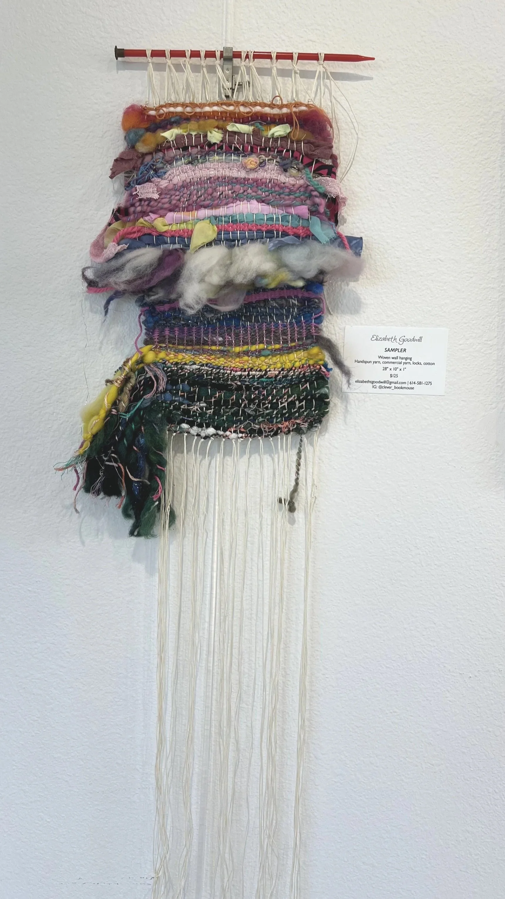 Wall hanging art piece made of woven yarns, fabrics, and loose fibers, attached to a horizontal pole with strings, displayed on a white wall.